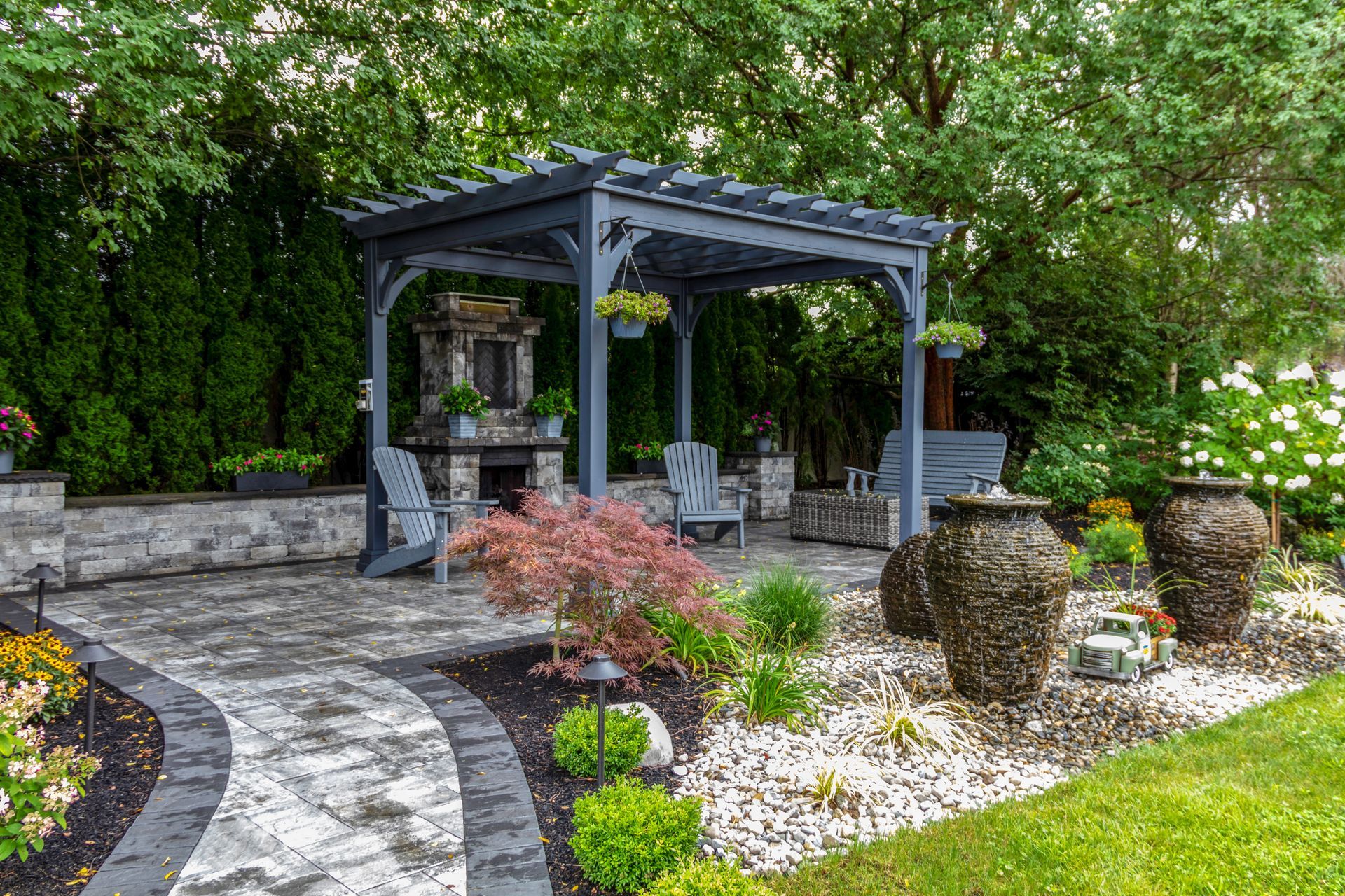 Stone patio with gray pergola, fireplace, chairs, water fountains, and landscaping.