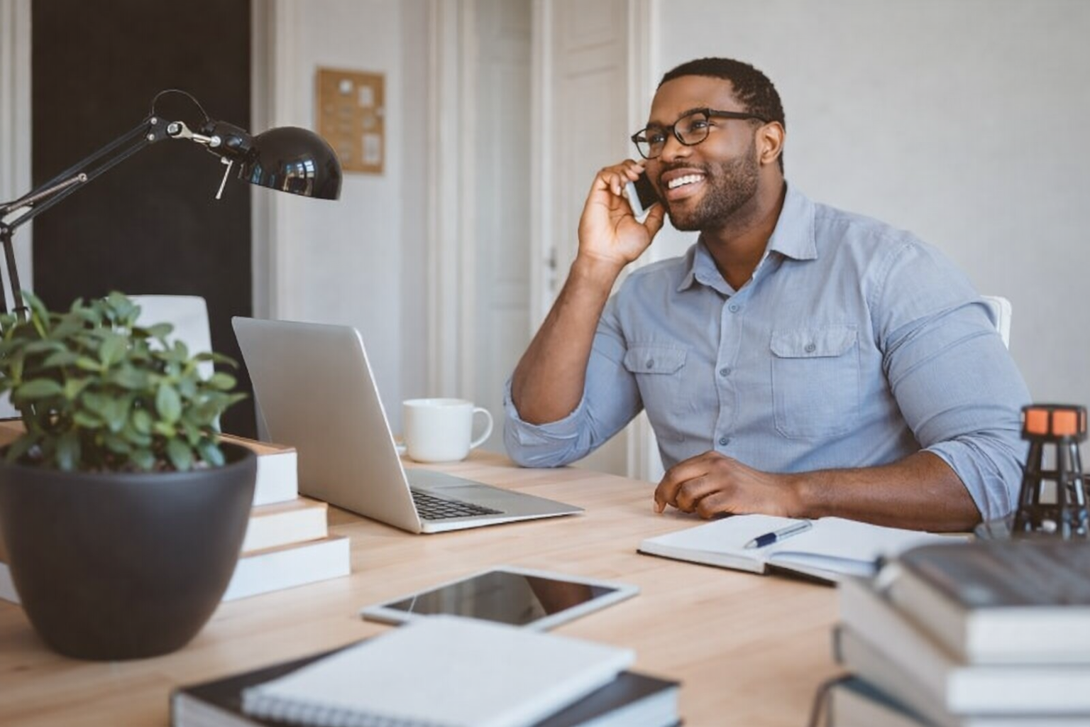 Man in blue shirt smiles while talking on a phone at a desk with laptop, plant, and notepad.