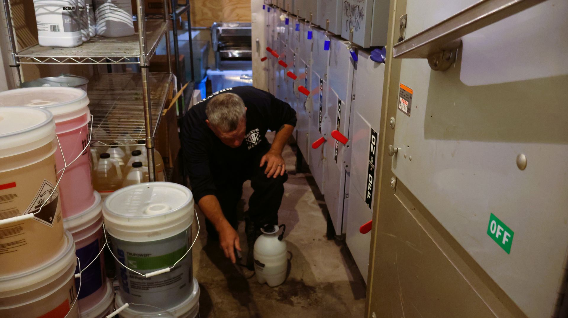 Person crouches on a boat cabin floor, handling a white canister beside stacked buckets and cabinets.