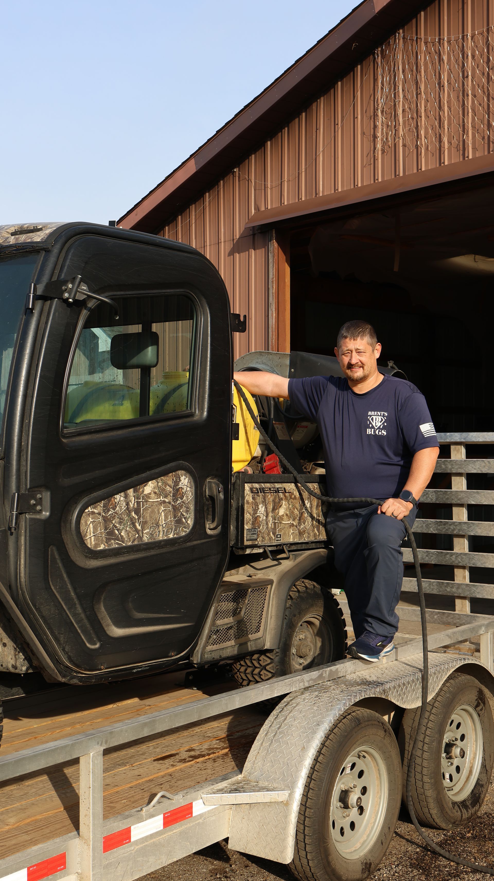 Man standing on a flatbed trailer beside a compact loader in front of a barn