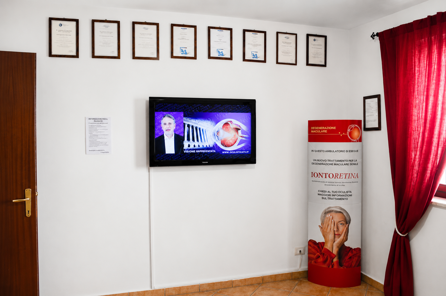 Televisore a parete in una stanza bianca con certificati incorniciati, una tenda rossa e un supporto per poster nell'angolo