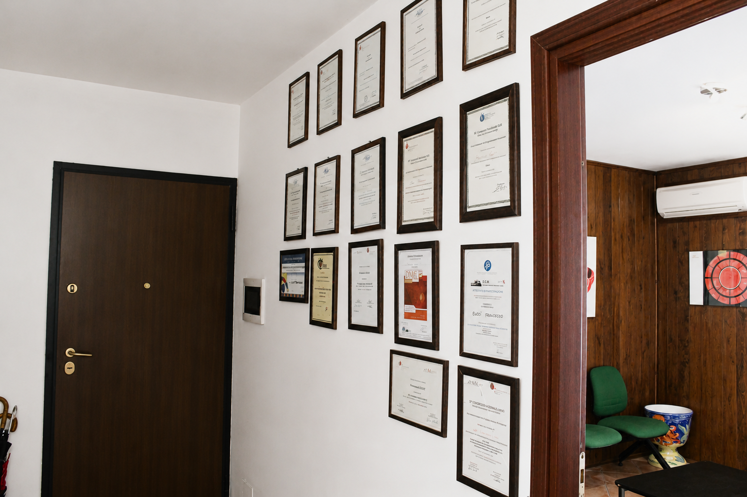 Corridoio con numerosi certificati incorniciati su una parete bianca, una porta scura e un'apertura che conduce a una stanza con sedie verdi.