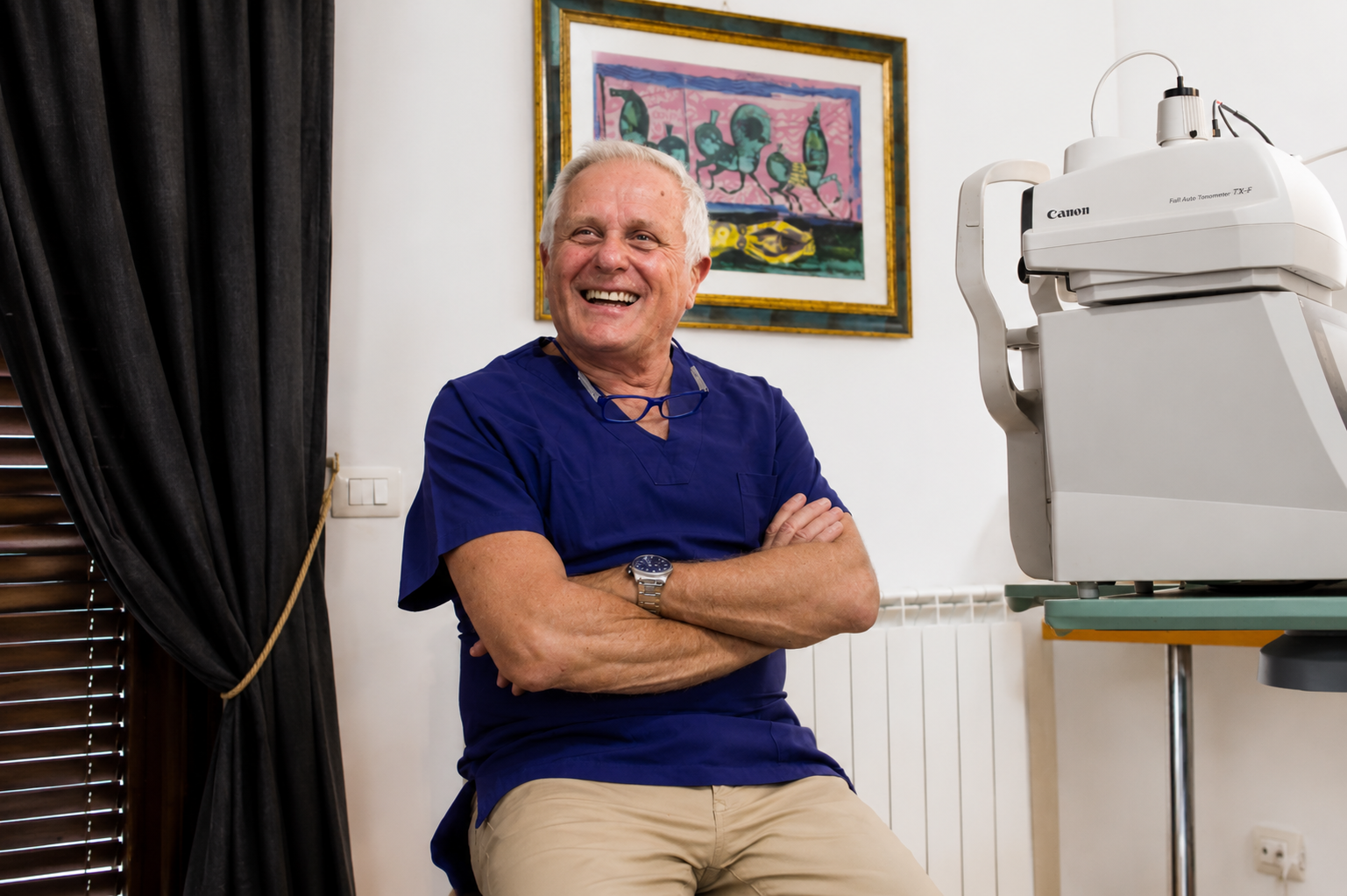 Un uomo sorridente con una camicia blu è seduto con le braccia incrociate accanto a delle apparecchiature dentistiche in una stanza luminosa.