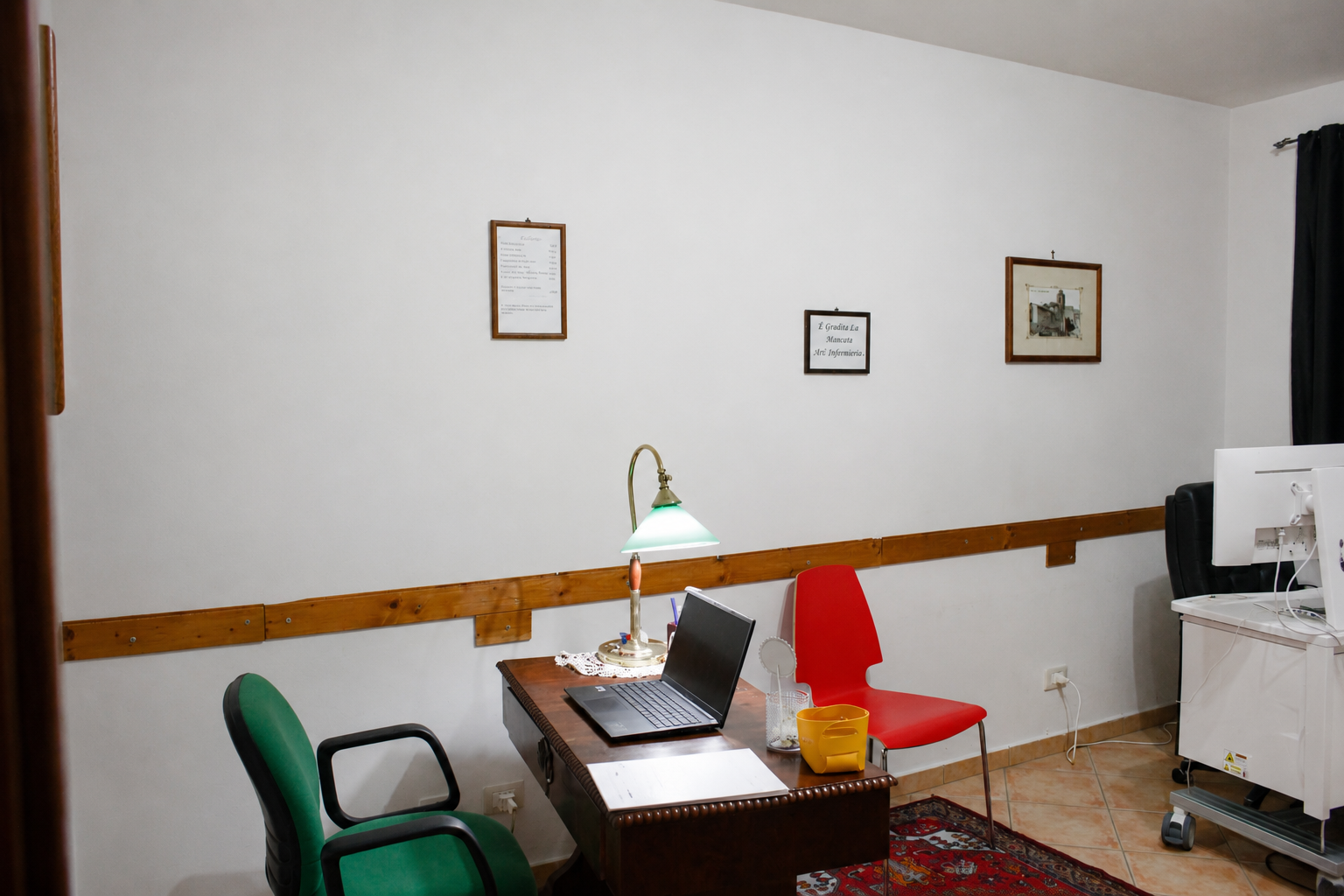 Piccolo ufficio con scrivania, computer portatile, lampada, sedia verde, sedia rossa e quadro incorniciato.