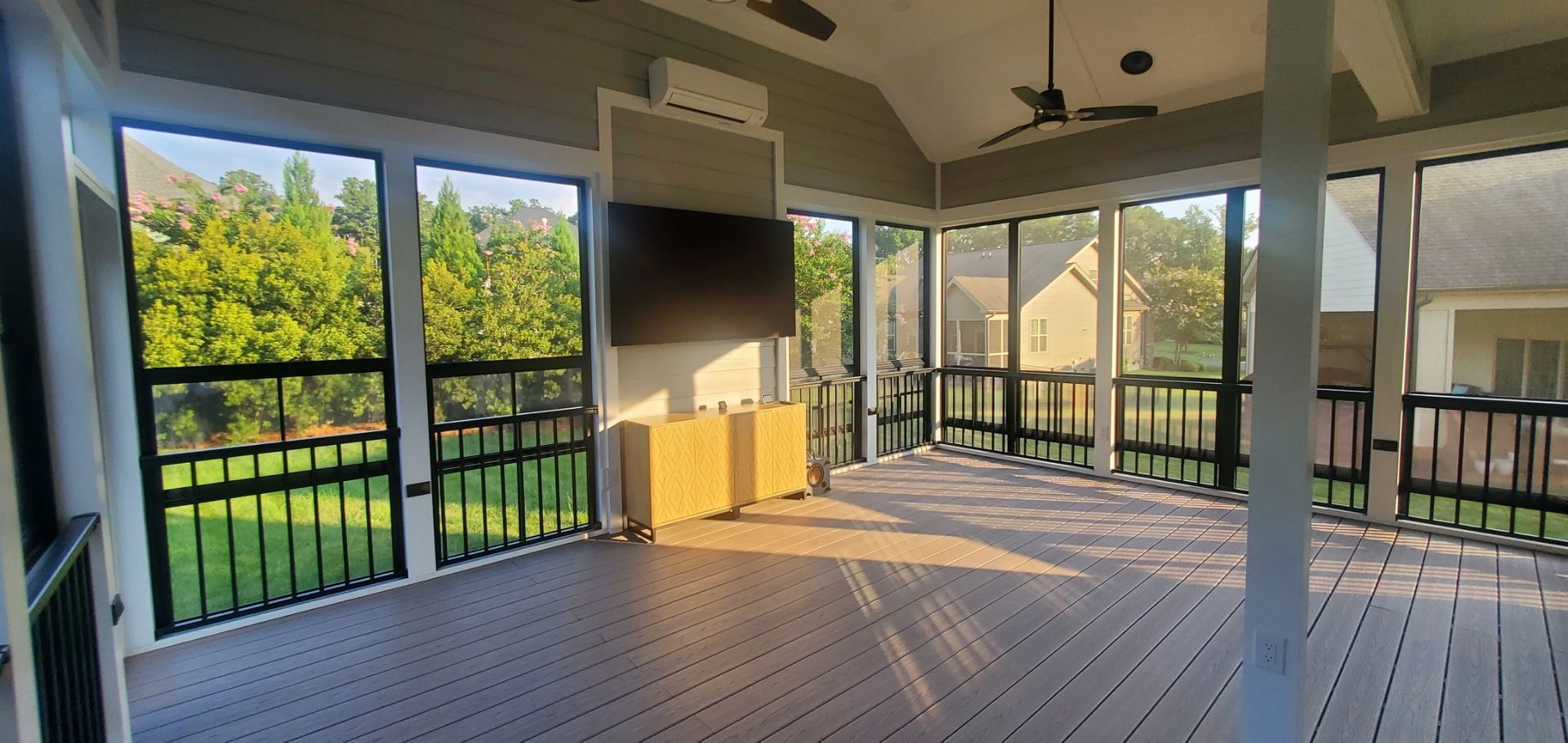Sunlit screened porch with white railings, wood floor, and views of green trees outside.