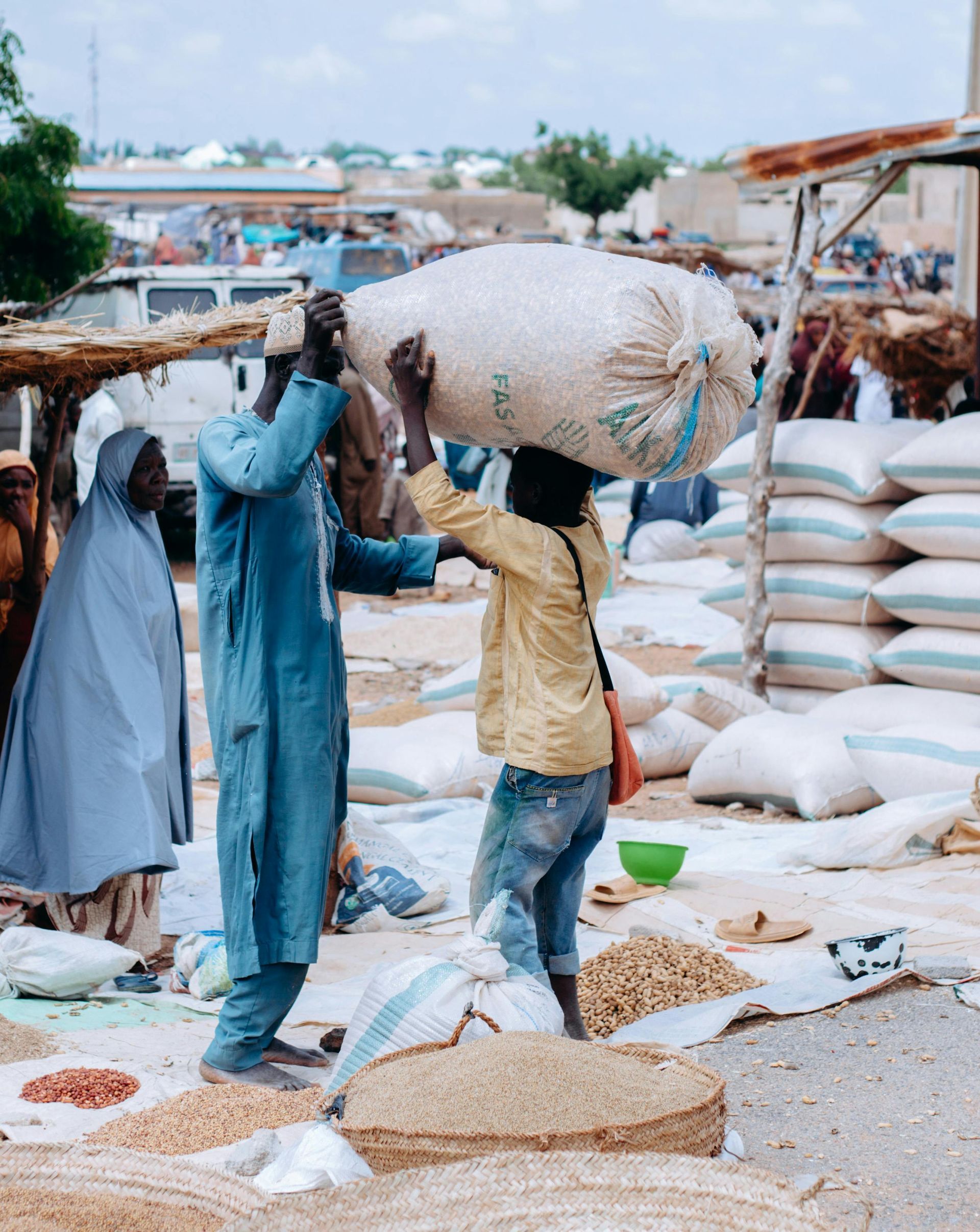 A man and a boy carrying a large sack of grain at an outdoor market. The market is bustling with activity.