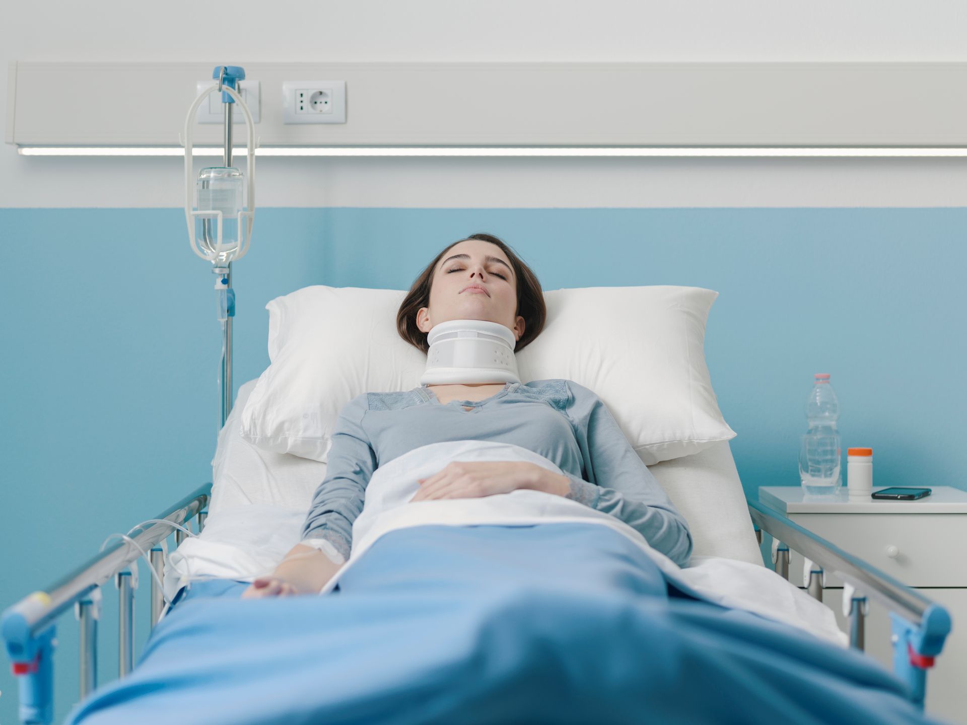 Woman in hospital bed wearing a neck brace; an IV drip is next to her, and a table with medication and a water bottle is nearby.