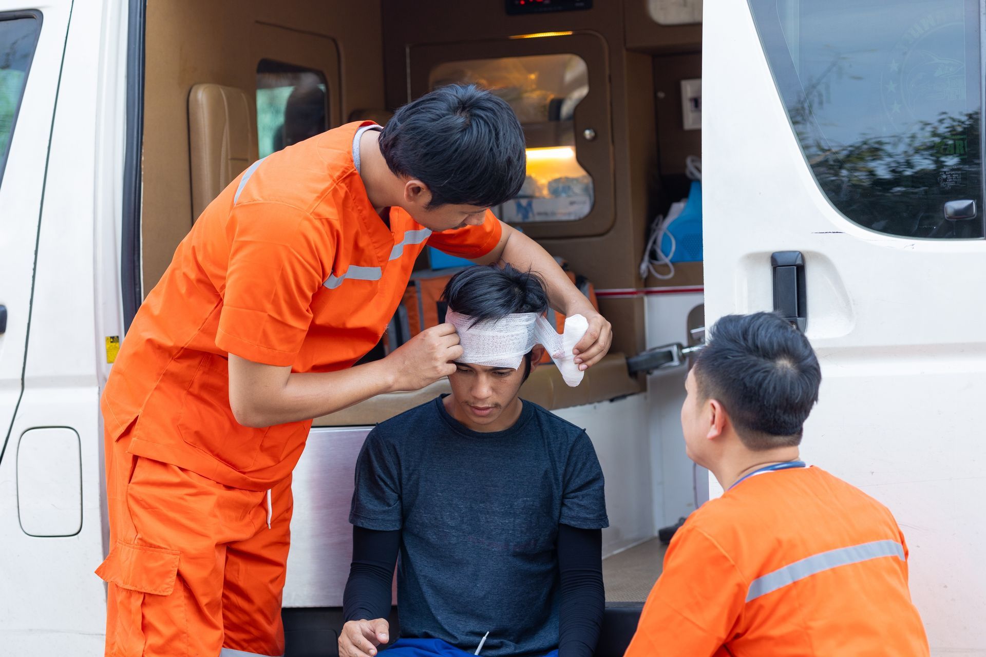 An EMT in an orange uniform bandages the head of a young man sitting in an ambulance, while another EMT watches.