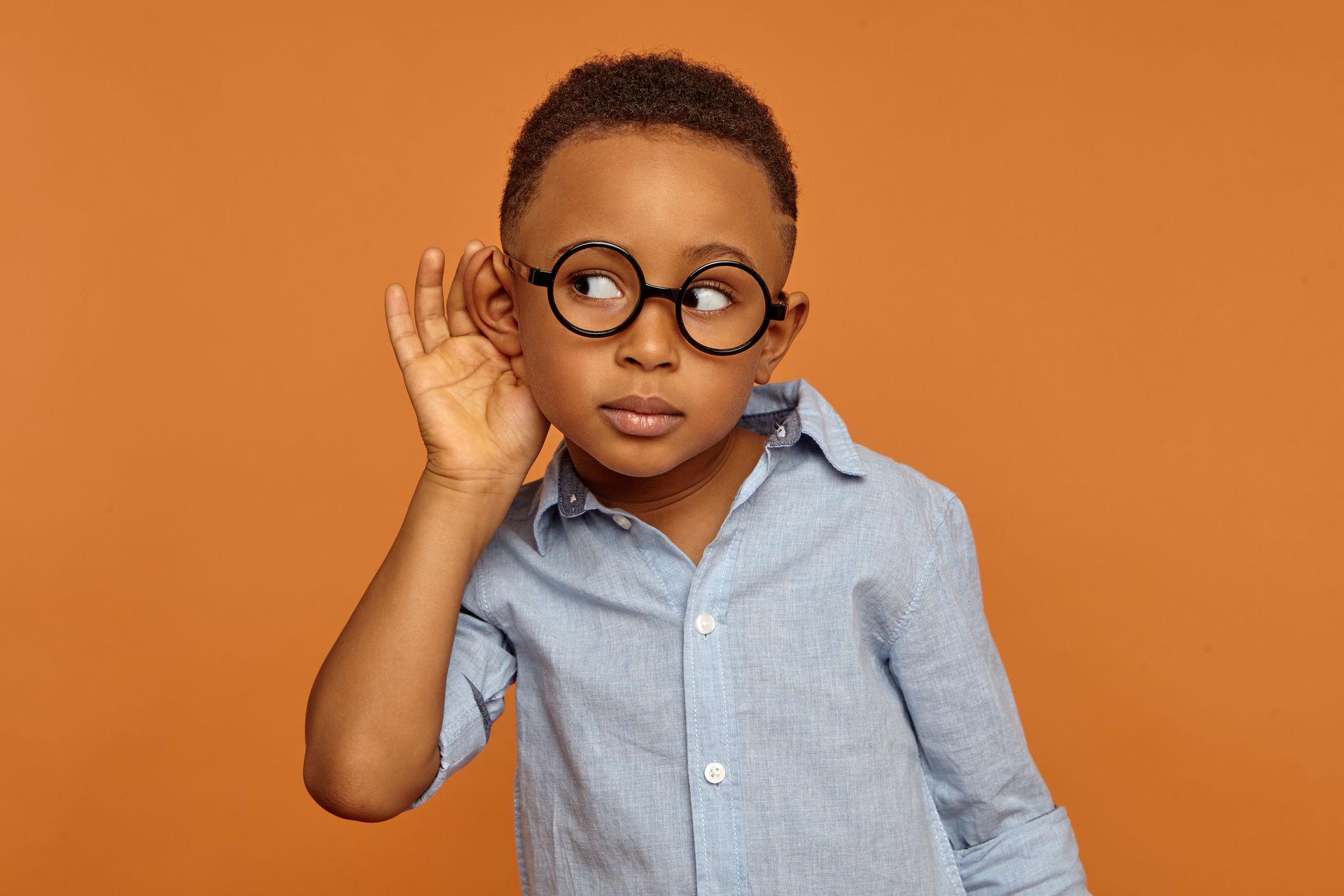A young boy wearing glasses is listening to something with his hand to his ear.