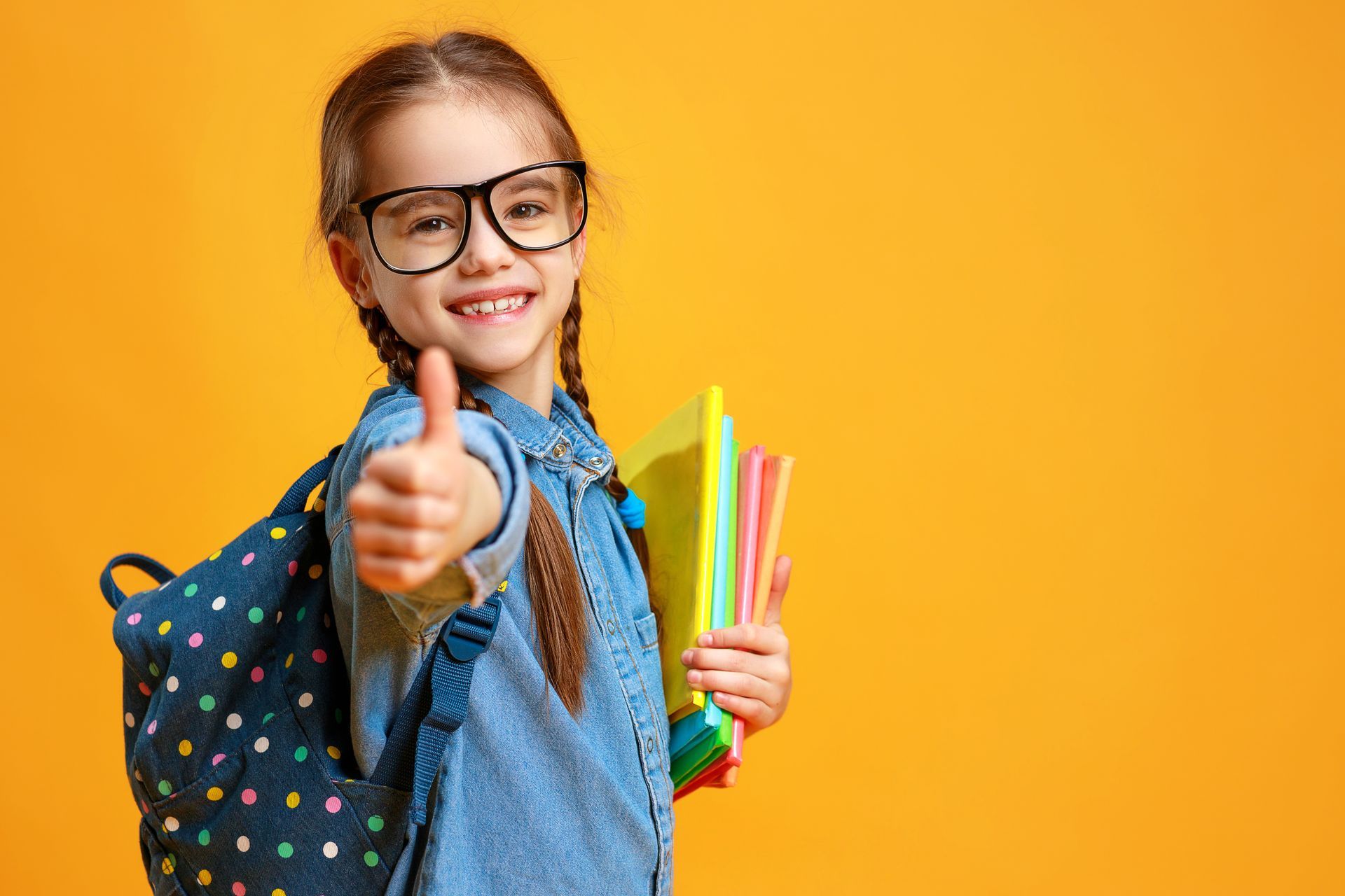 A little girl with glasses and a backpack is giving a thumbs up.