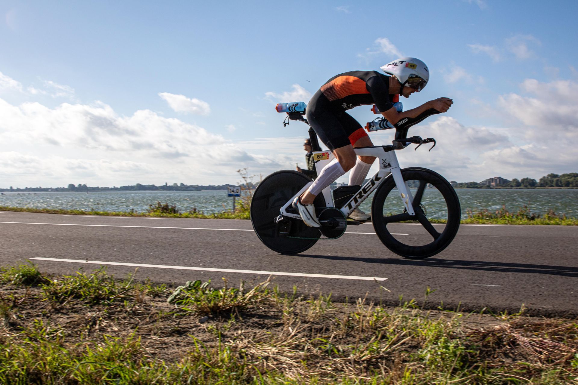 Triathlete on white Tri bike with Tailwind Wheels in aero position on a road. Wearing helmet, black jersey, black shorts, coastal setting.