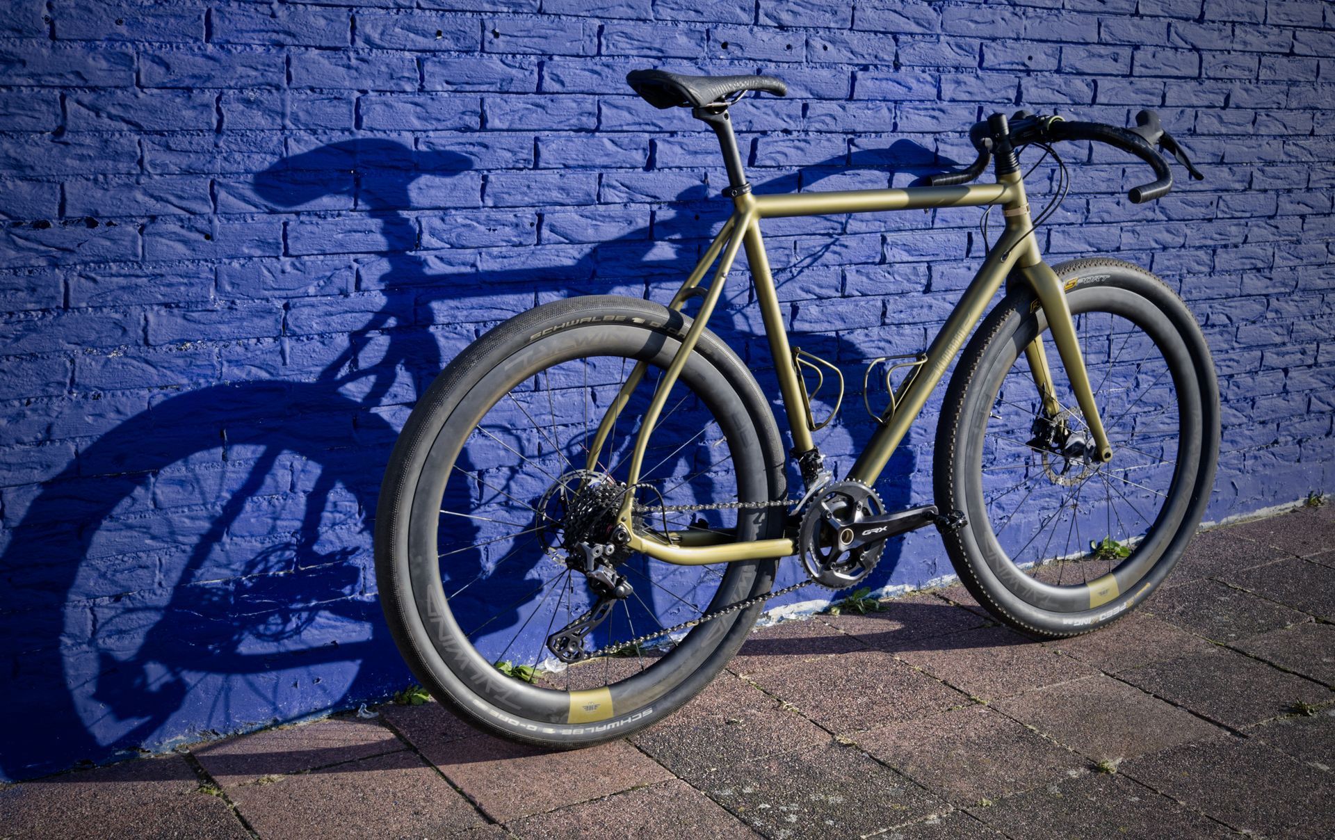 A Klaassen multipurpose gravel bike with carbon Tailwind wheels is parked in front of a blue brick wall.