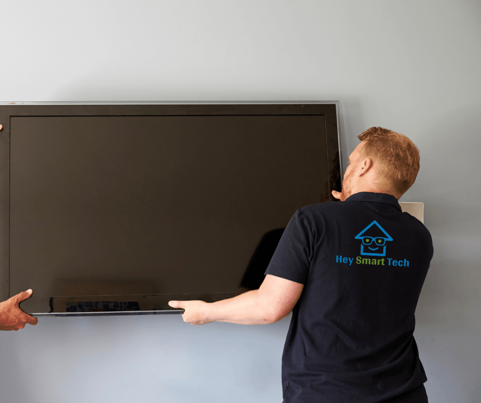Two people mounting a large black TV on a gray wall; one person is wearing a "Hey Smart Tech" t-shirt.