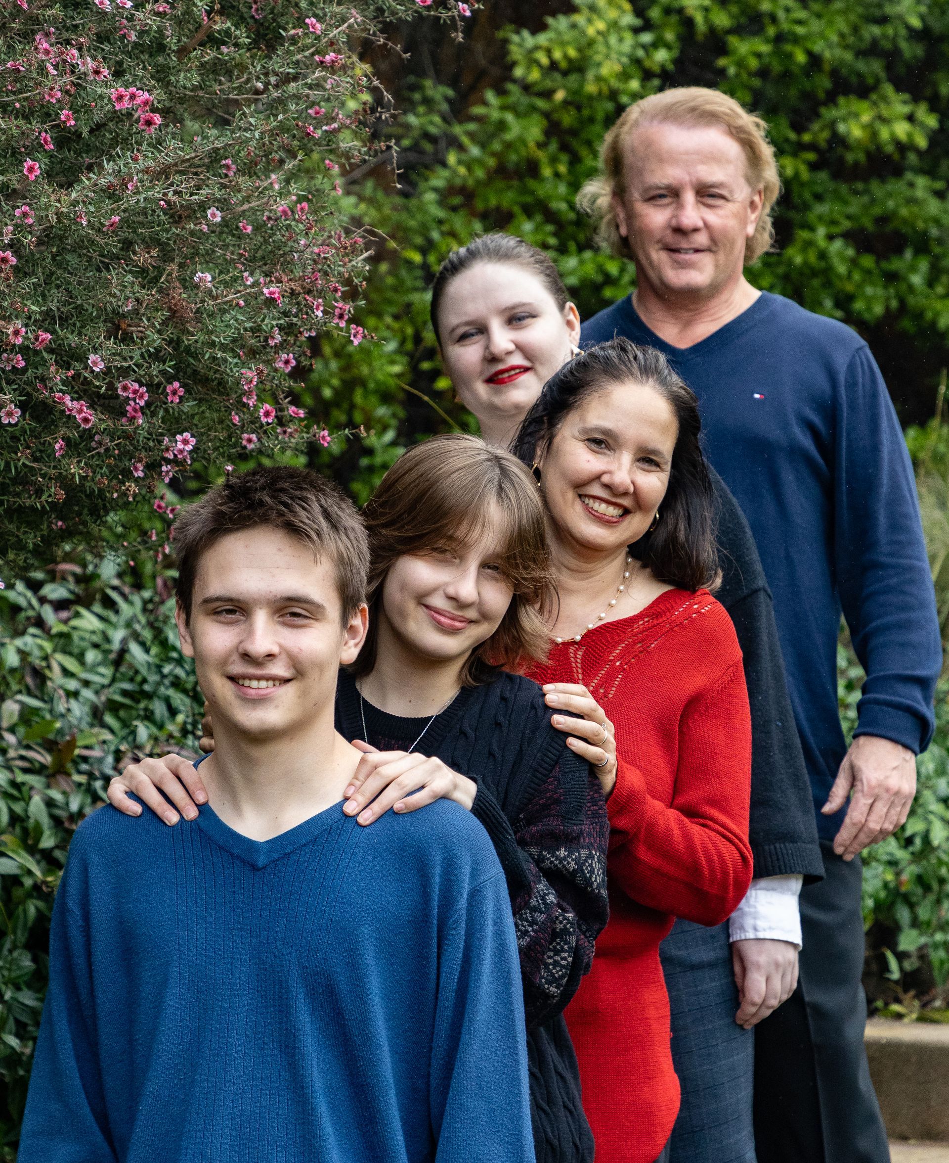 Family of five smiling, standing in a line outdoors; green foliage background.