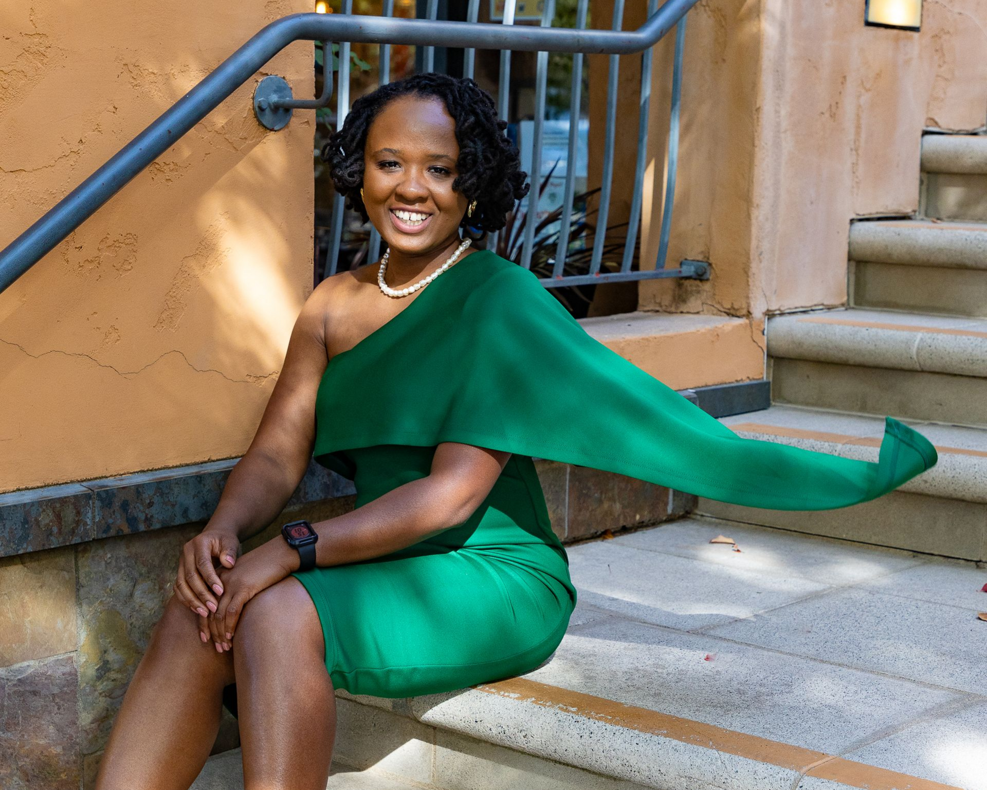 Woman in a green one-shoulder dress sits on stone steps, smiling. A hand rests on her thigh.