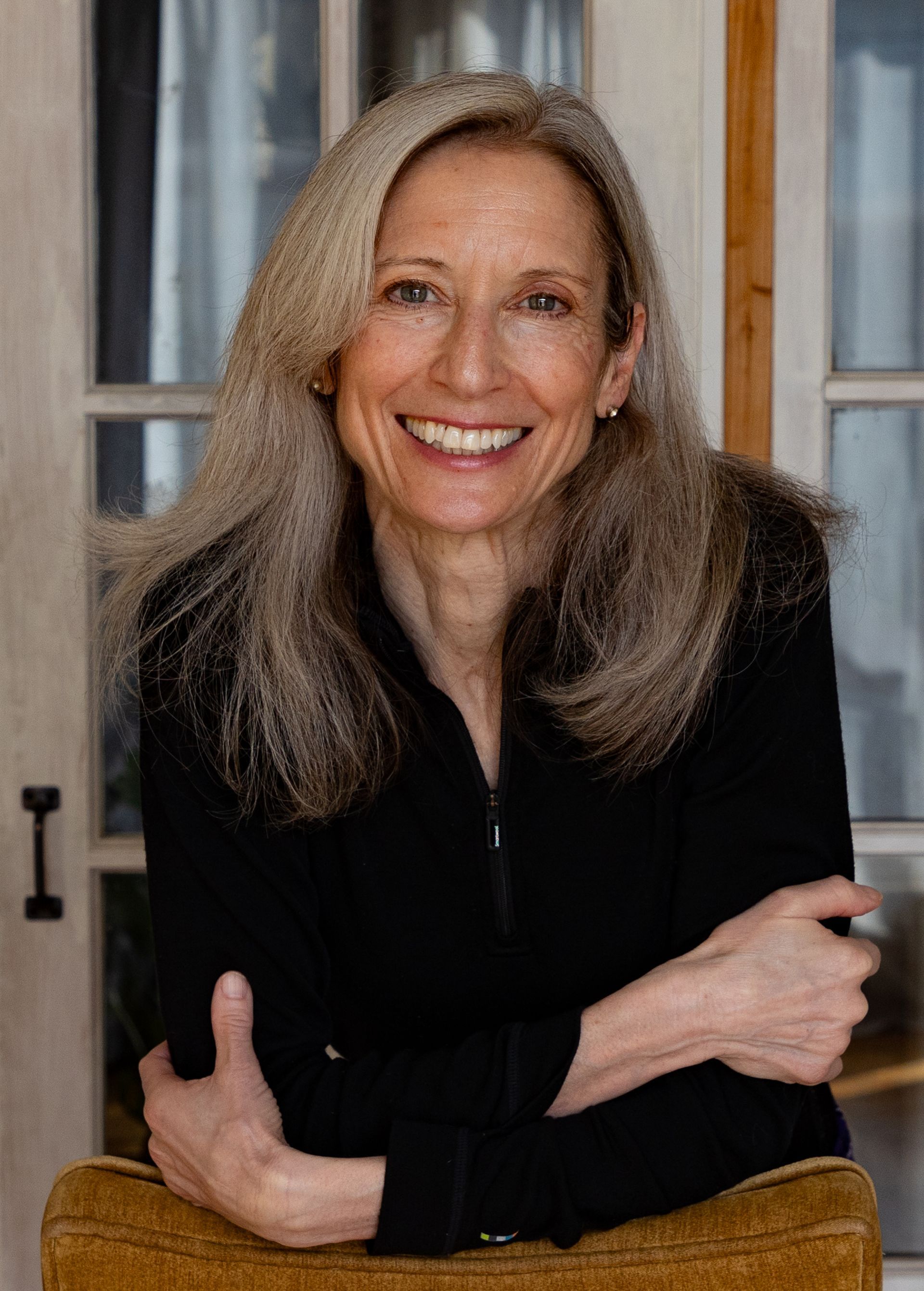 Woman with long gray hair smiling, leaning on a yellow chair, wearing black top, in a room with white doors.