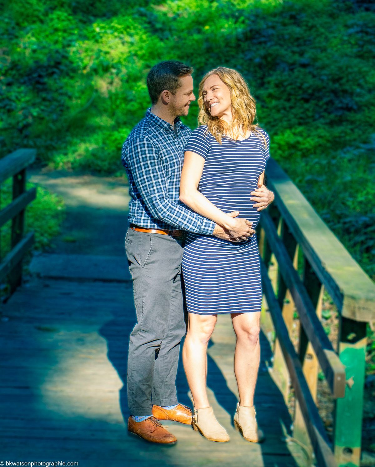 Couple hugging on a wooden bridge in a green park. Woman wearing striped dress, man in plaid shirt.