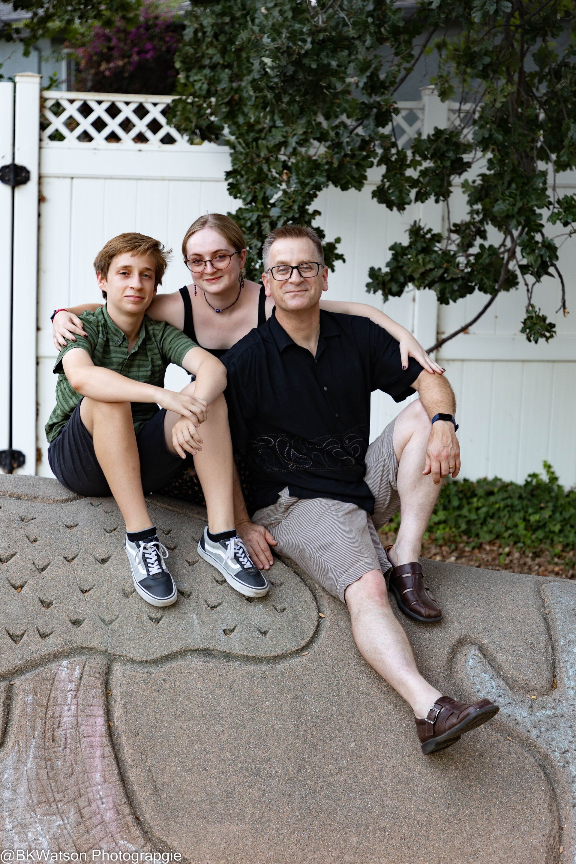 Family sitting together outdoors on a stone structure. Man in black shirt, shorts, and two teens smiling.