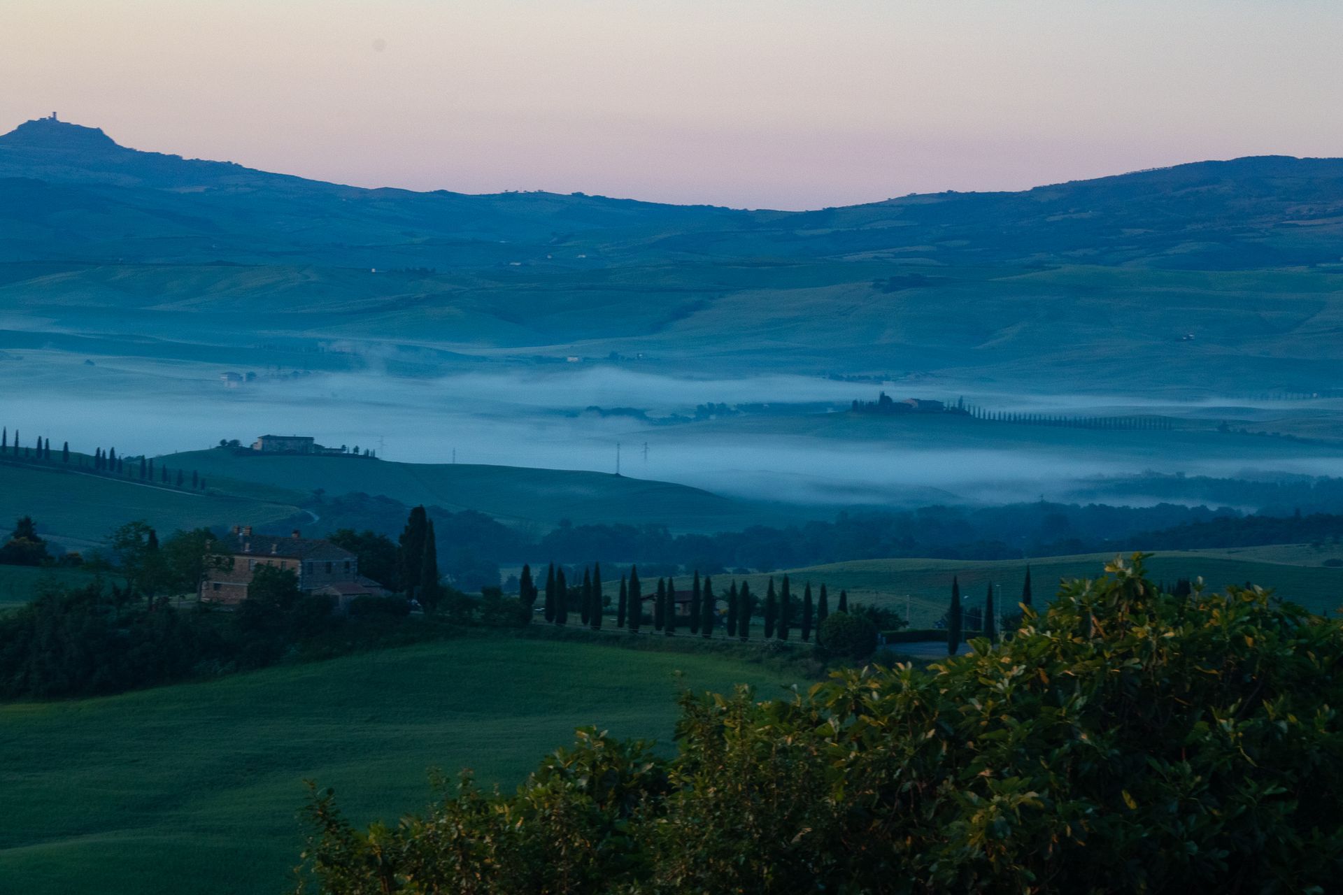 Rolling green hills of Tuscany at dawn, with misty valleys and a cypress-lined road.