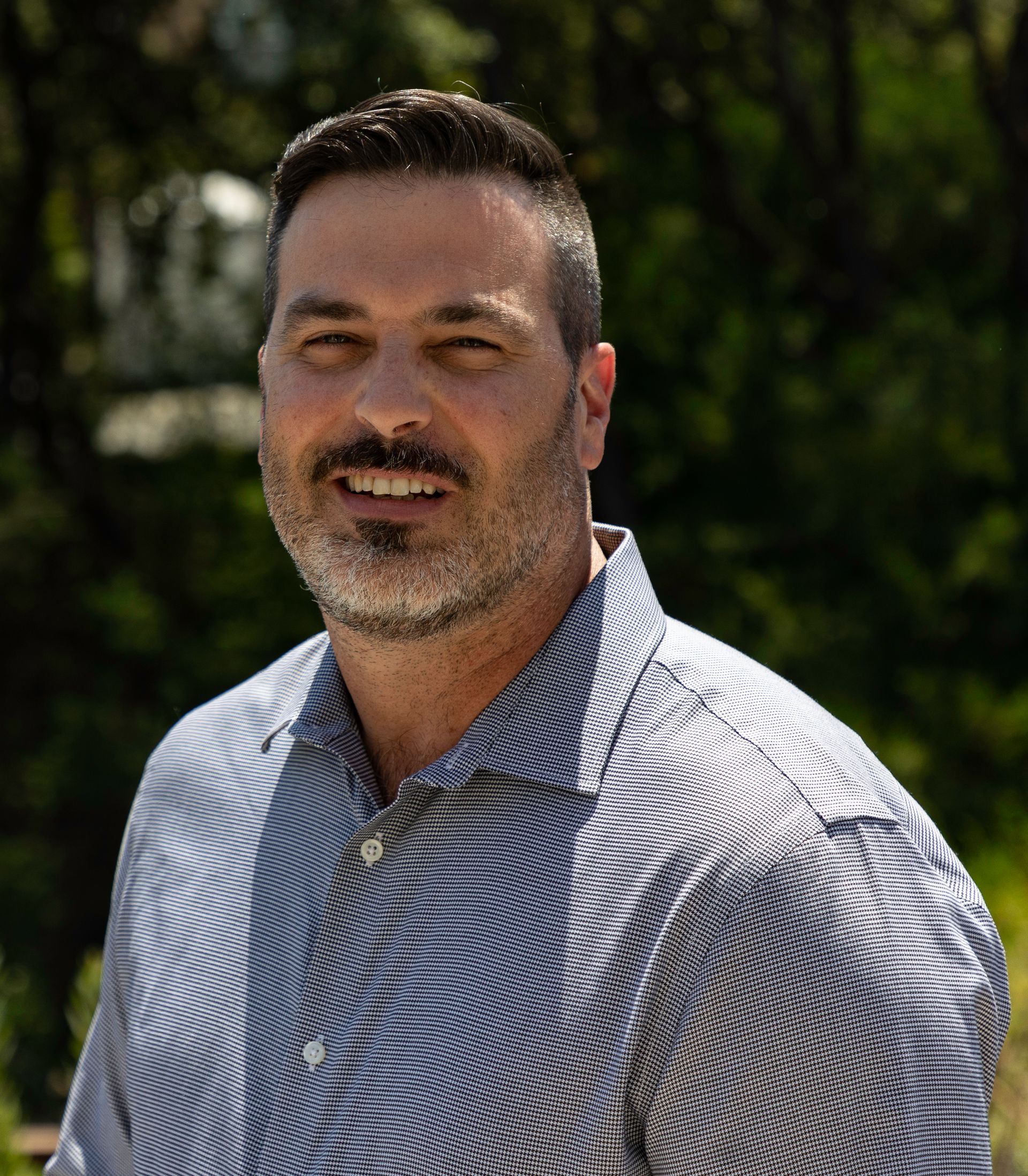 Man with short dark hair, graying beard, smiling in blue striped shirt outdoors.