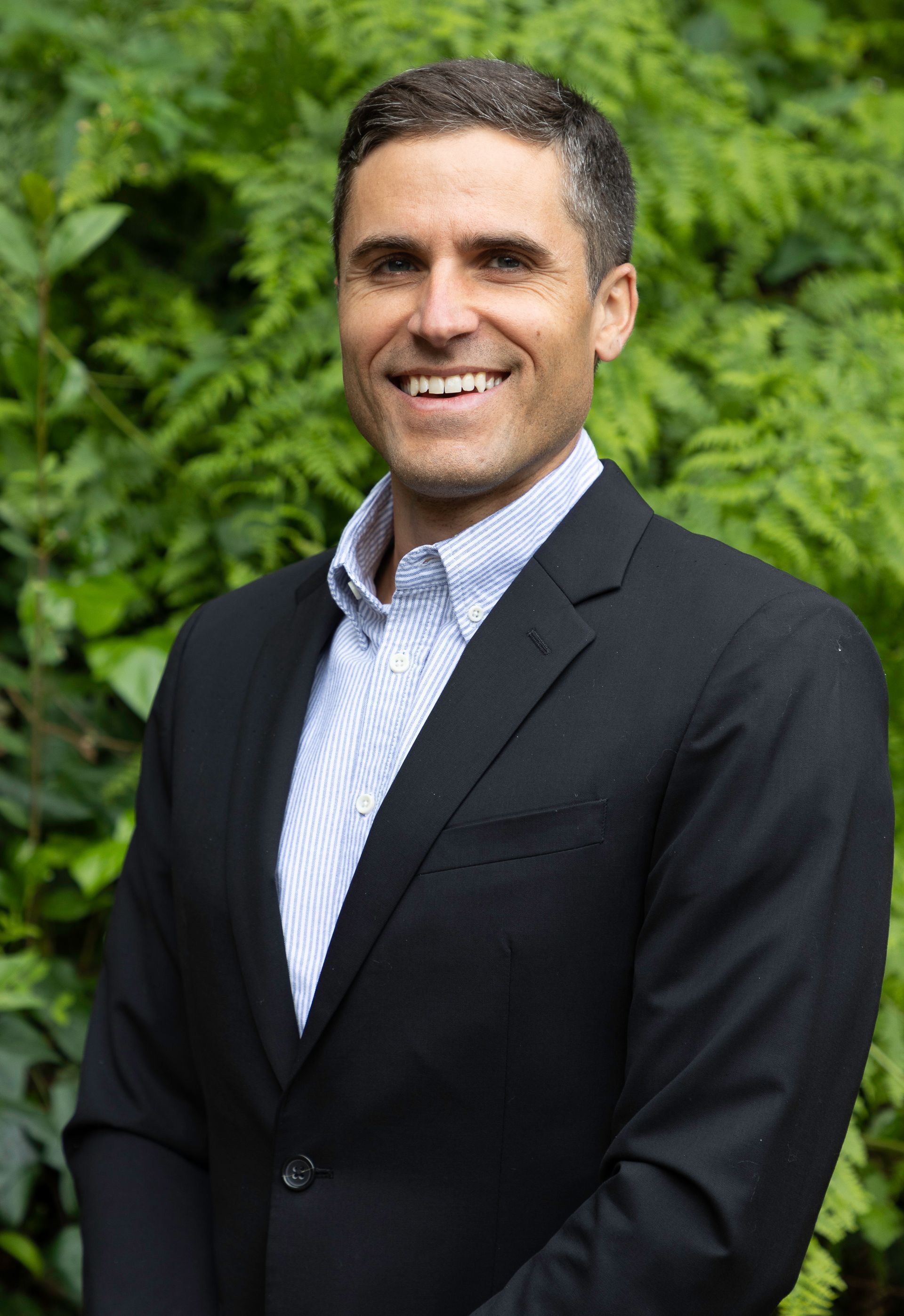 Man in a black blazer and blue shirt smiles in front of green foliage.