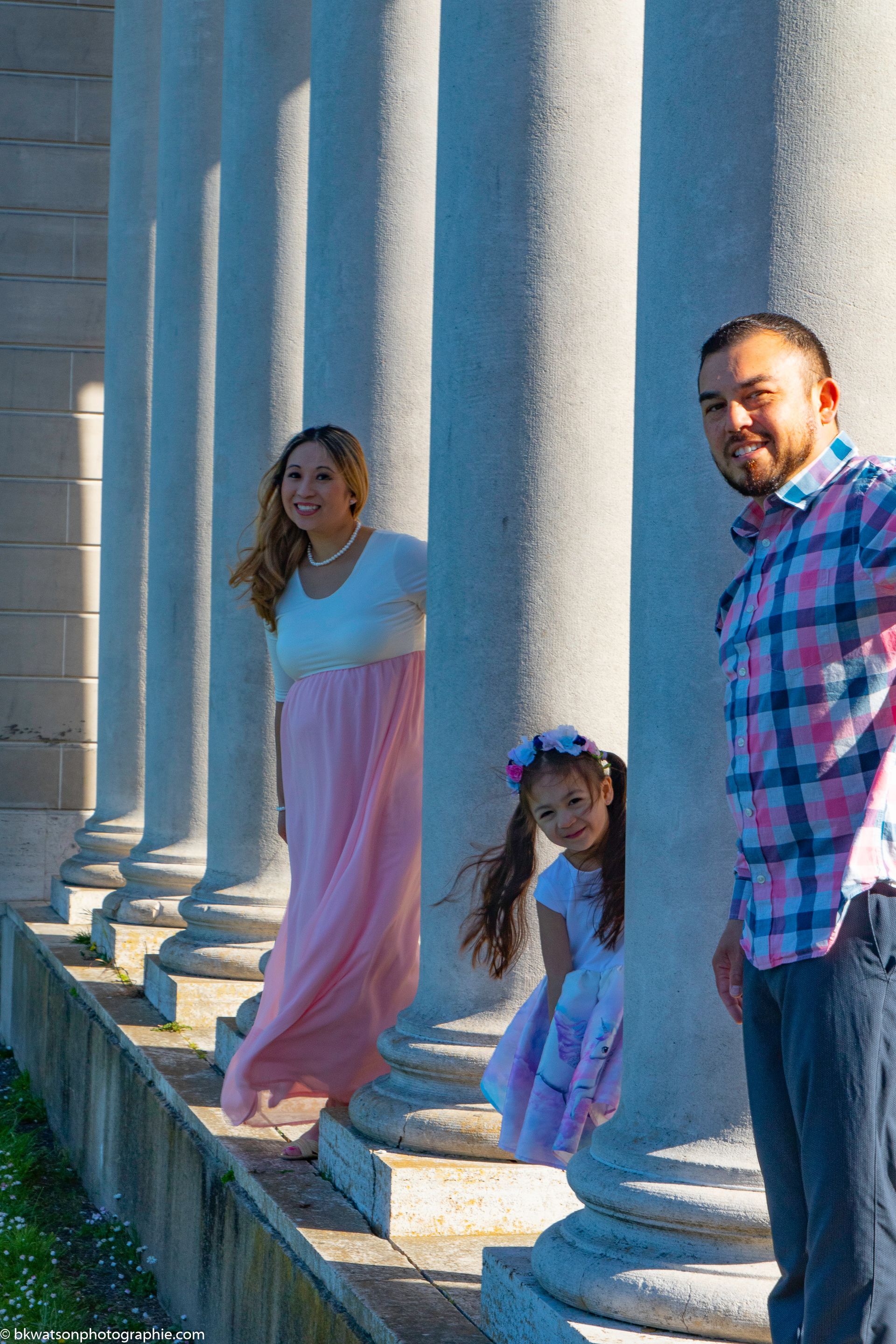 Family posing between white columns: woman in pink dress smiles, child peers out, man smiles.