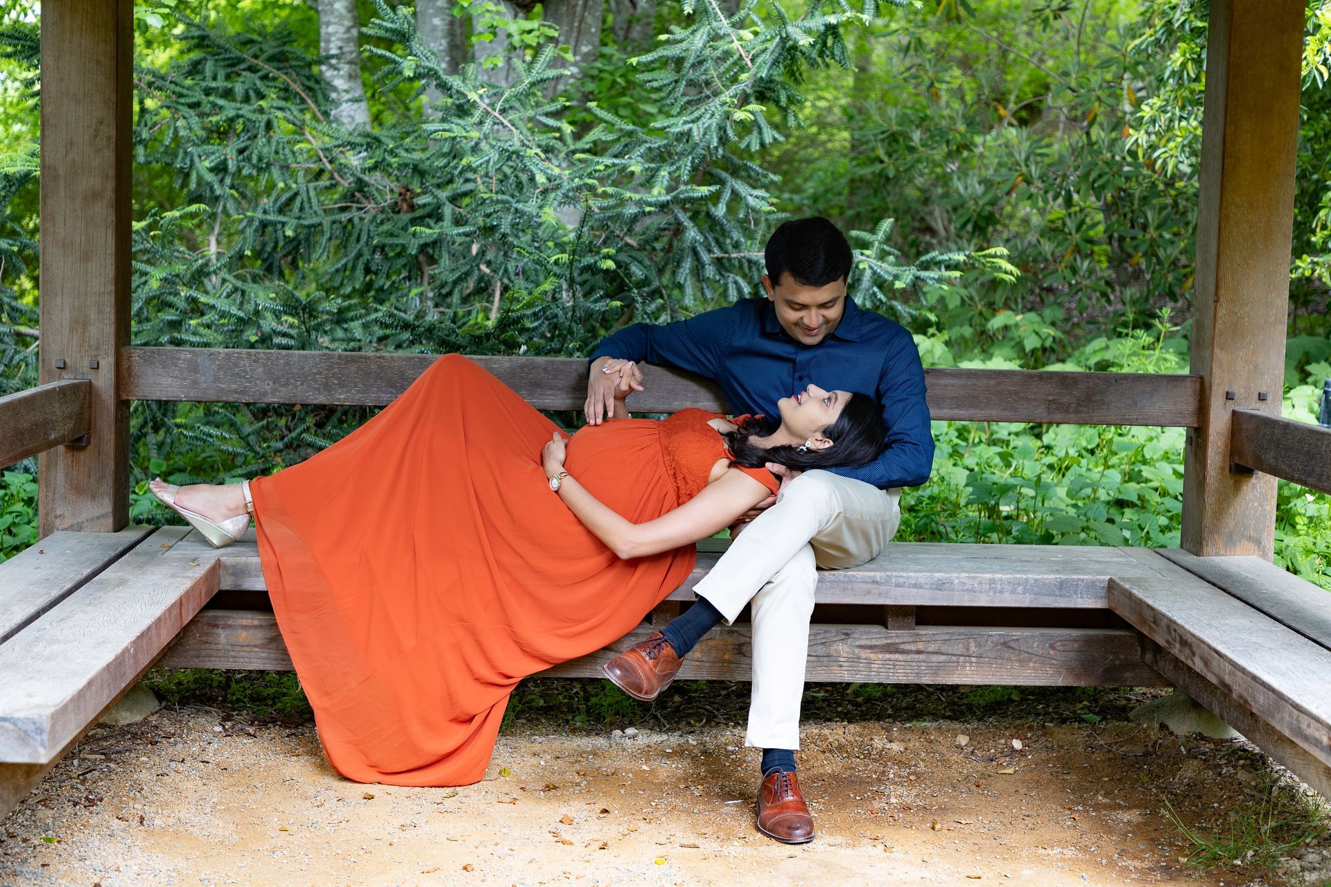Couple in maternity shoot. Woman in orange dress resting head on man's lap on wooden bench, in forest.