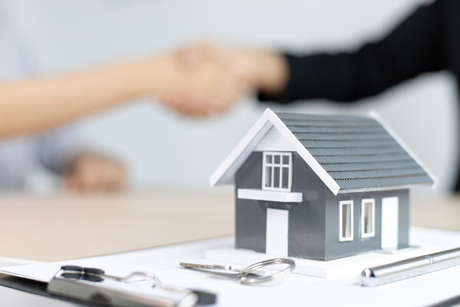 Miniature house, keys, pen, and document with blurred handshake in the background symbolizing a real estate deal.