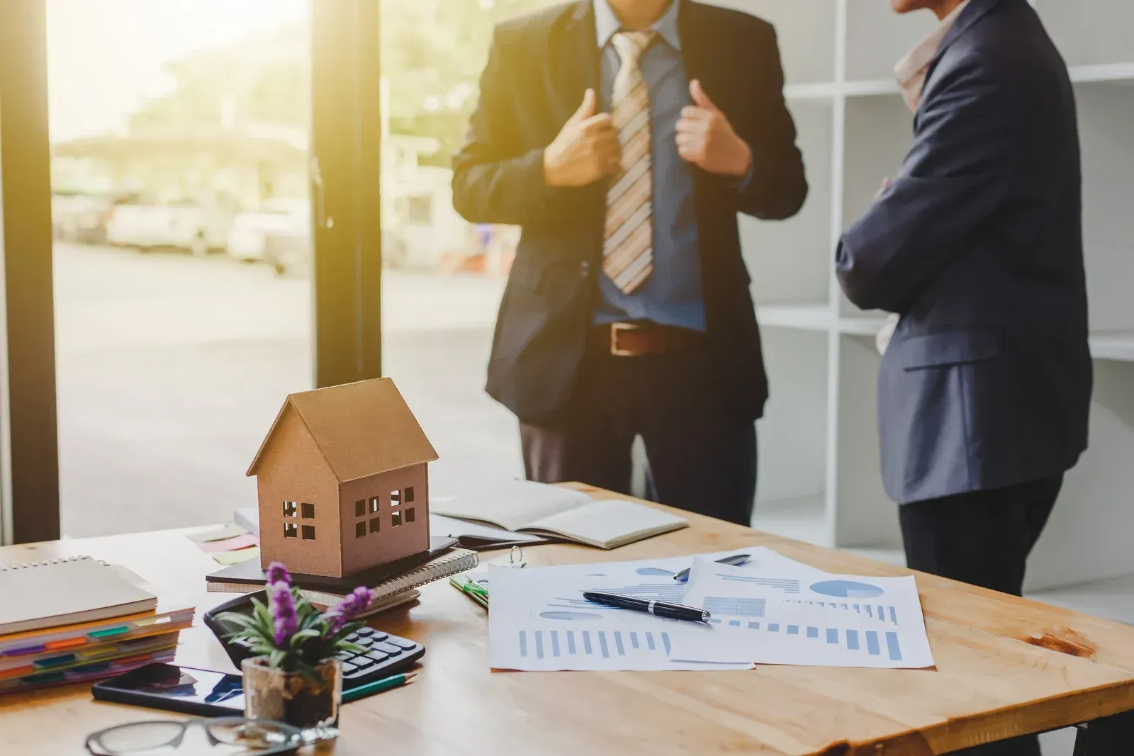 Two businesspeople discussing paperwork at a desk with a model house and financial documents.