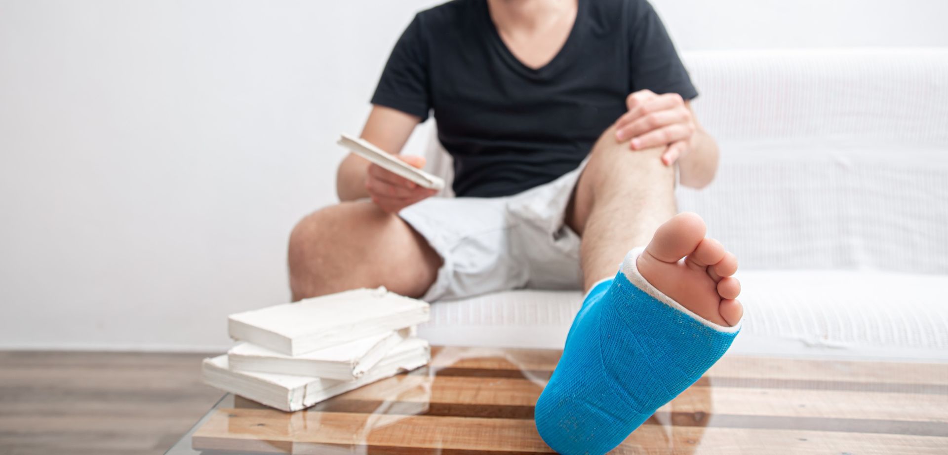 Person with a broken leg sitting on a couch, holding a notepad, with a blue cast and books nearby