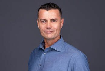 Man in a blue button-up shirt posing against a dark gray background, slight smile