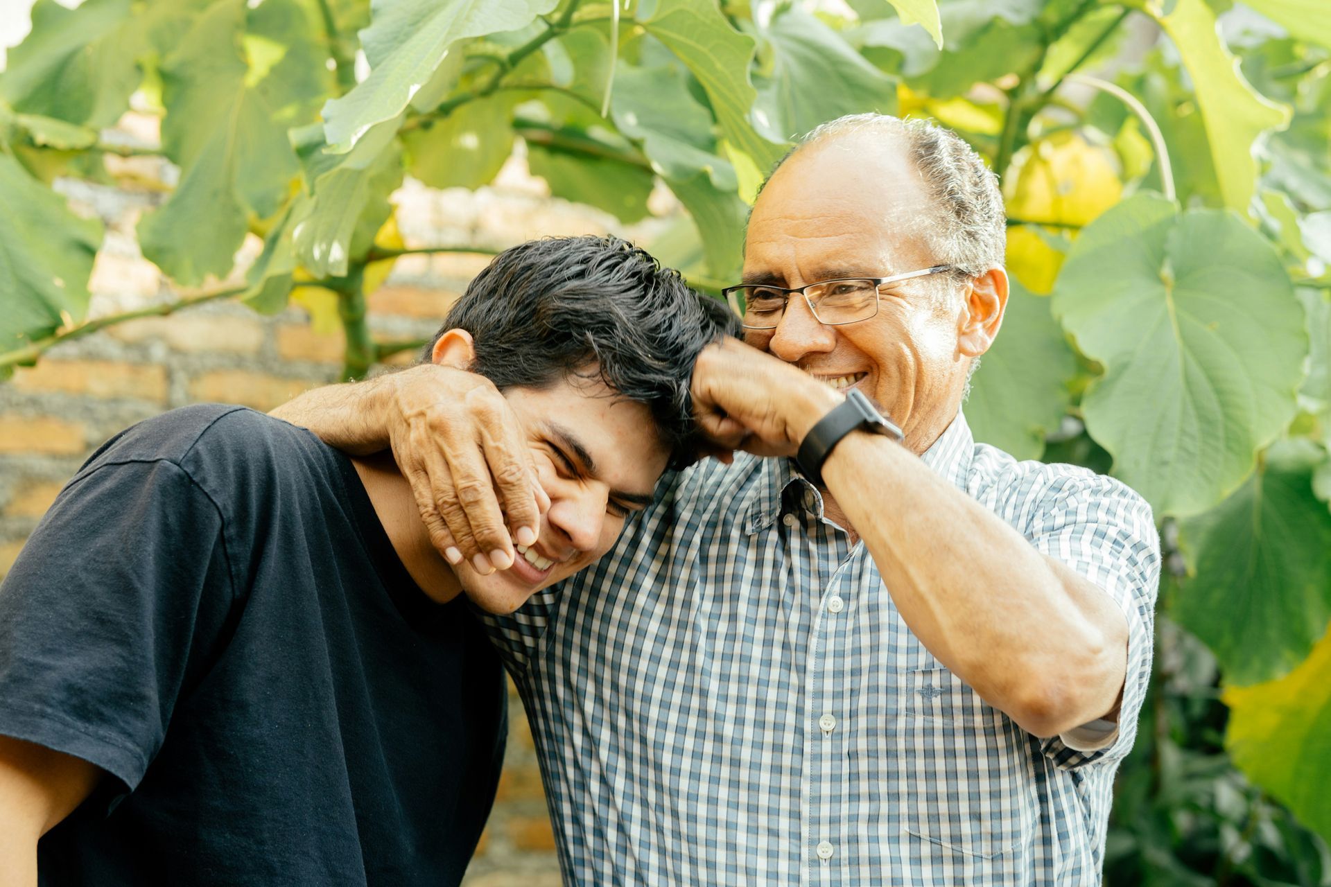 Two smiling people lean together in a leafy garden, one covering the other's eyes.