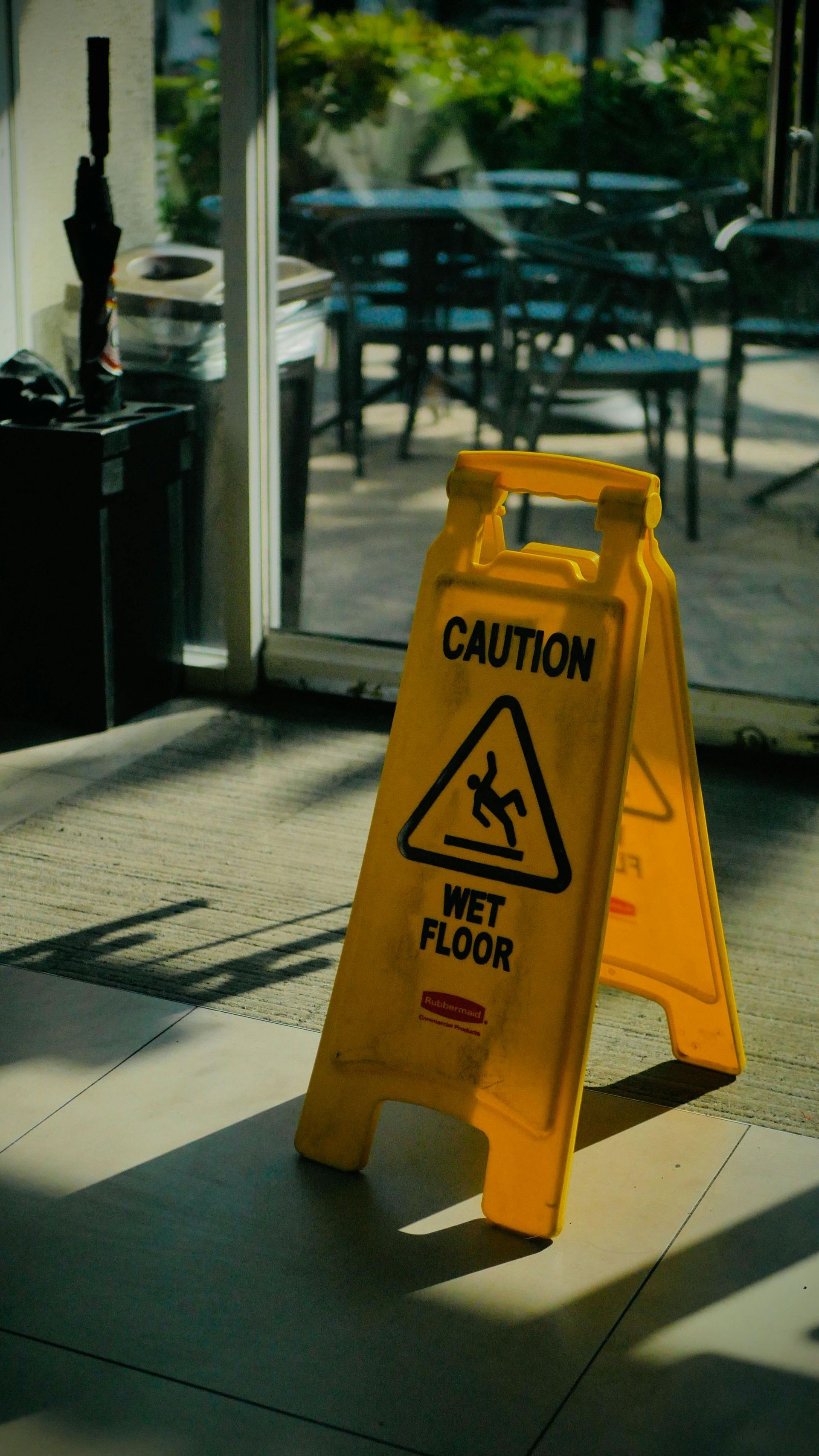 Yellow caution sign reading “Wet Floor” in front of a glass door, with patio tables in the background.