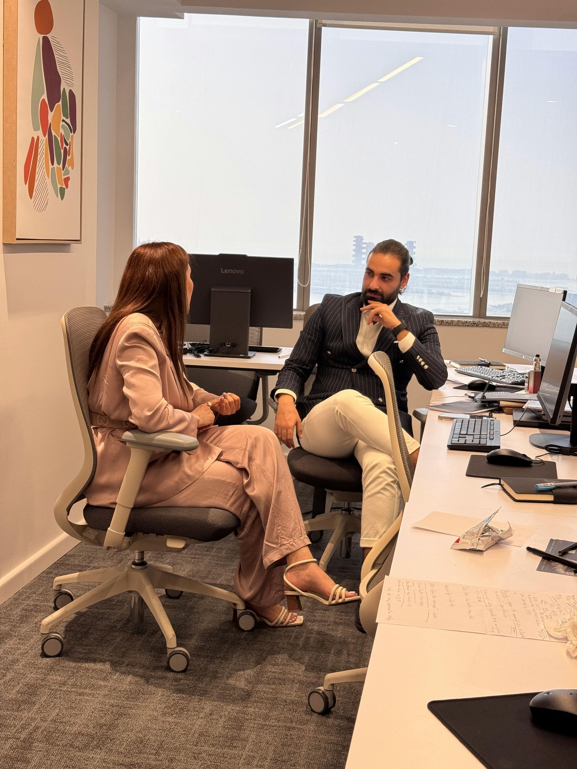 Two people talking in a bright office, seated at desks with computers and papers.