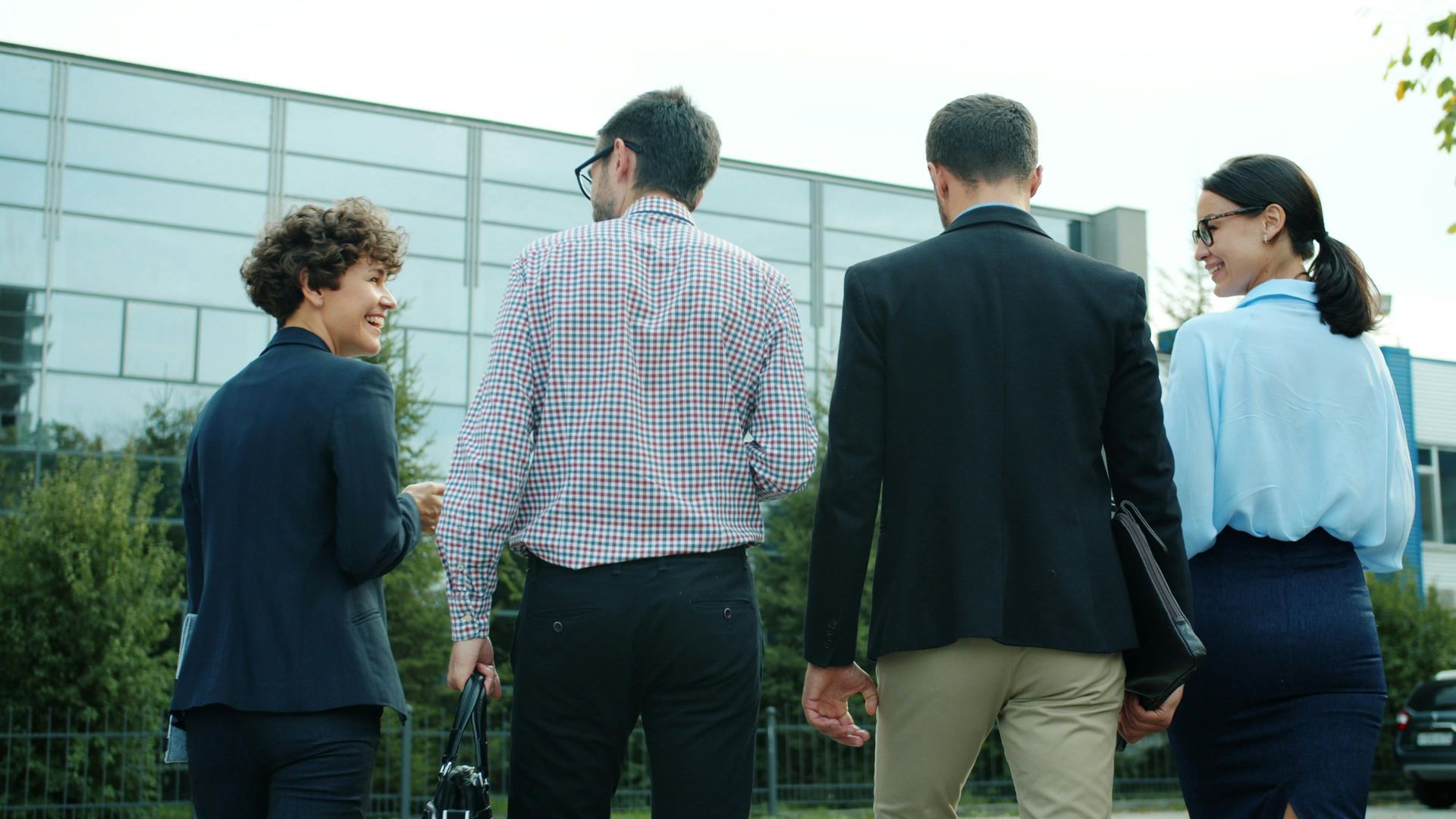 Four coworkers stand outdoors, seen from behind, facing a modern glass building.