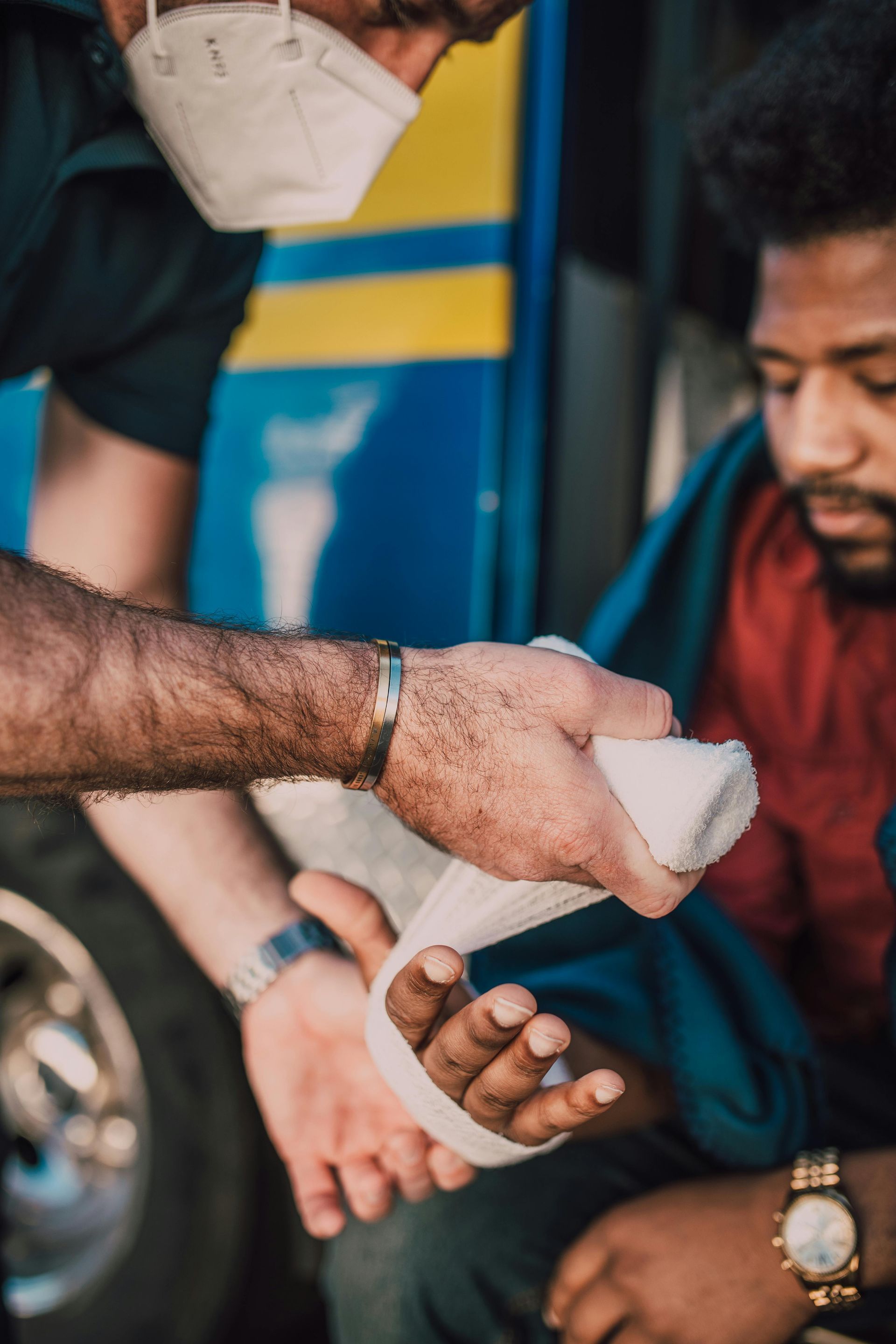 Person wrapping a white bandage around another person’s wrist while seated outdoors.