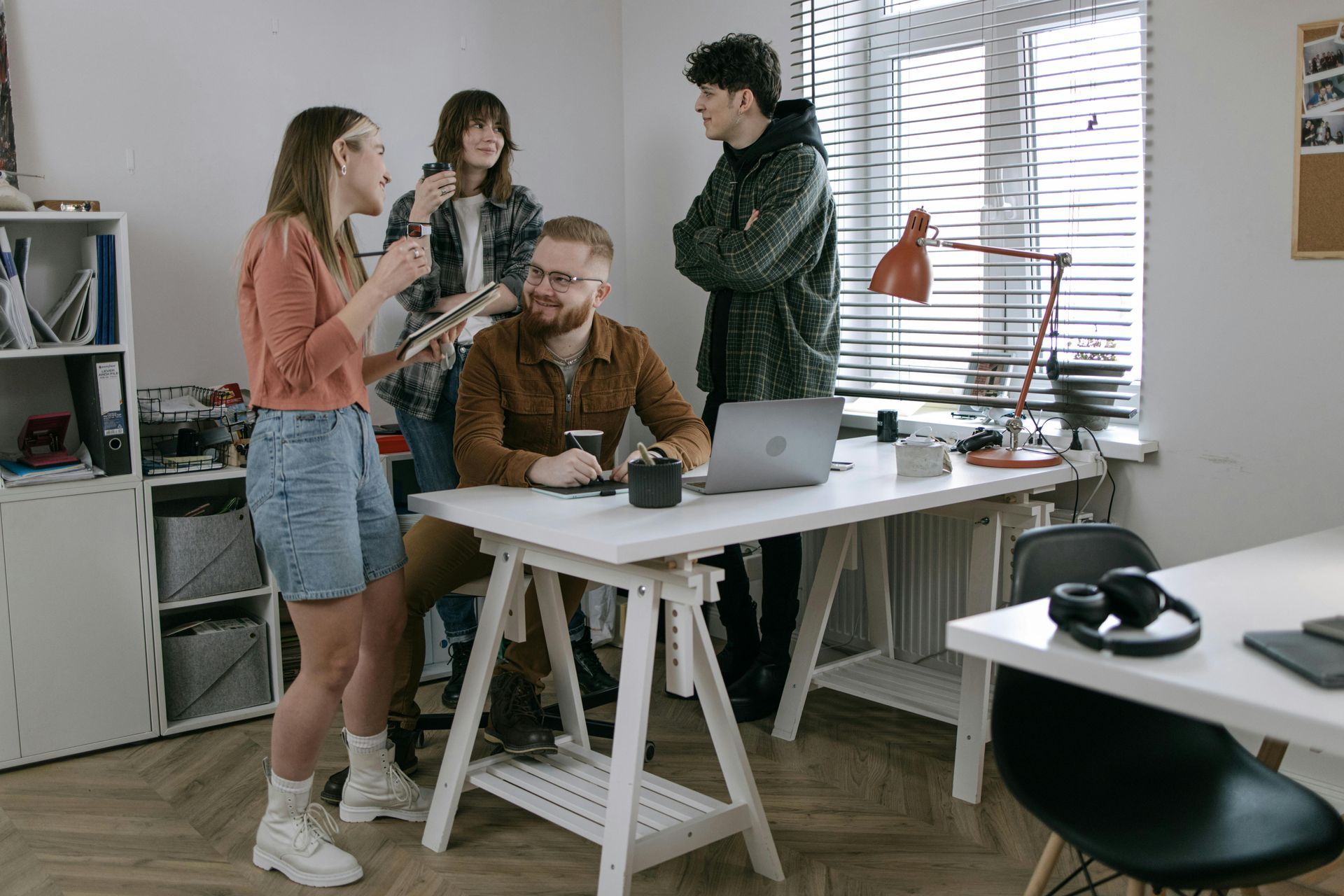 Four coworkers gathered around a desk in a bright office, discussing a laptop screen.