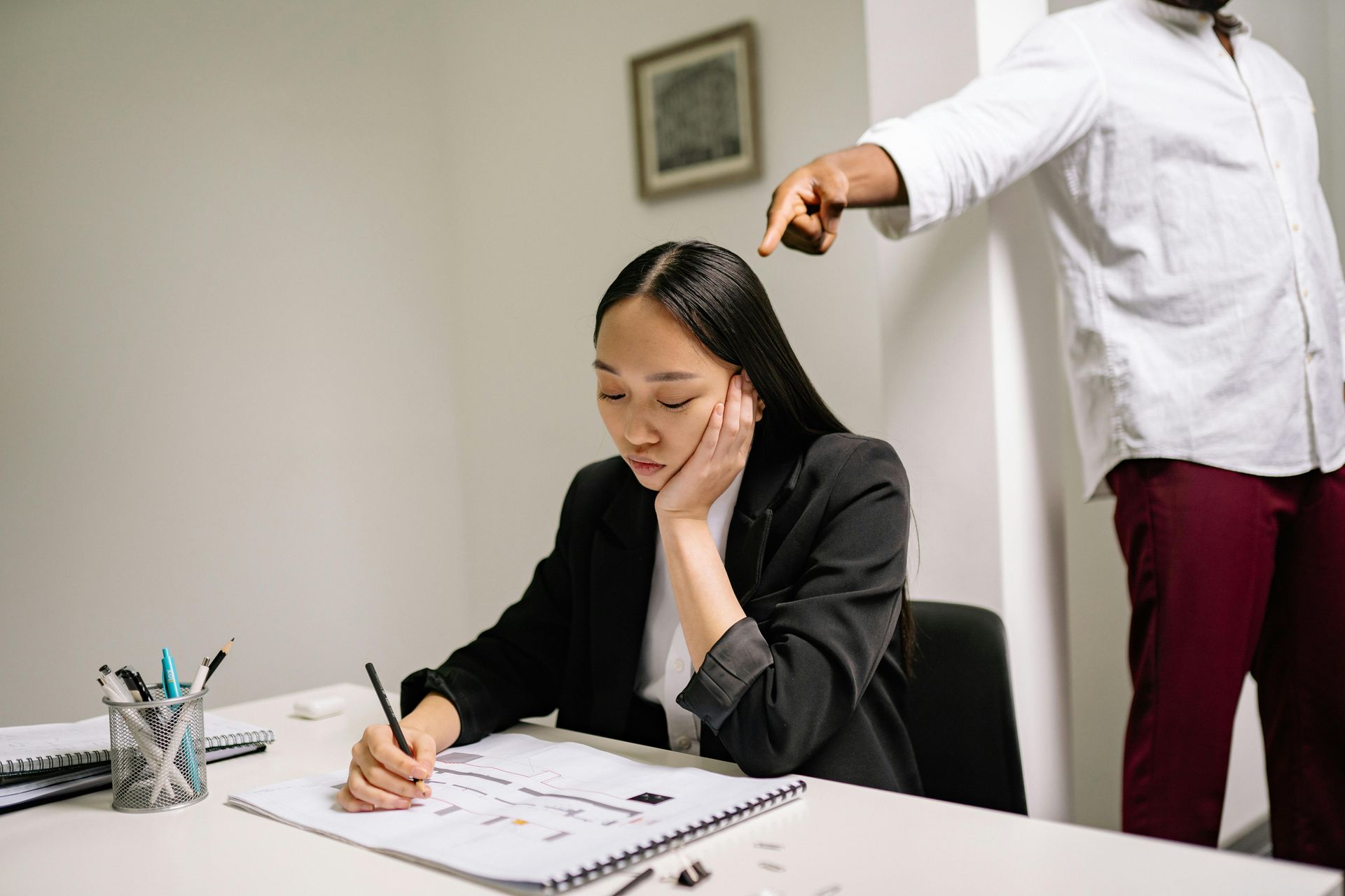 Woman sitting at a desk while a man reaches toward her head in an office, with notebooks and pens nearby