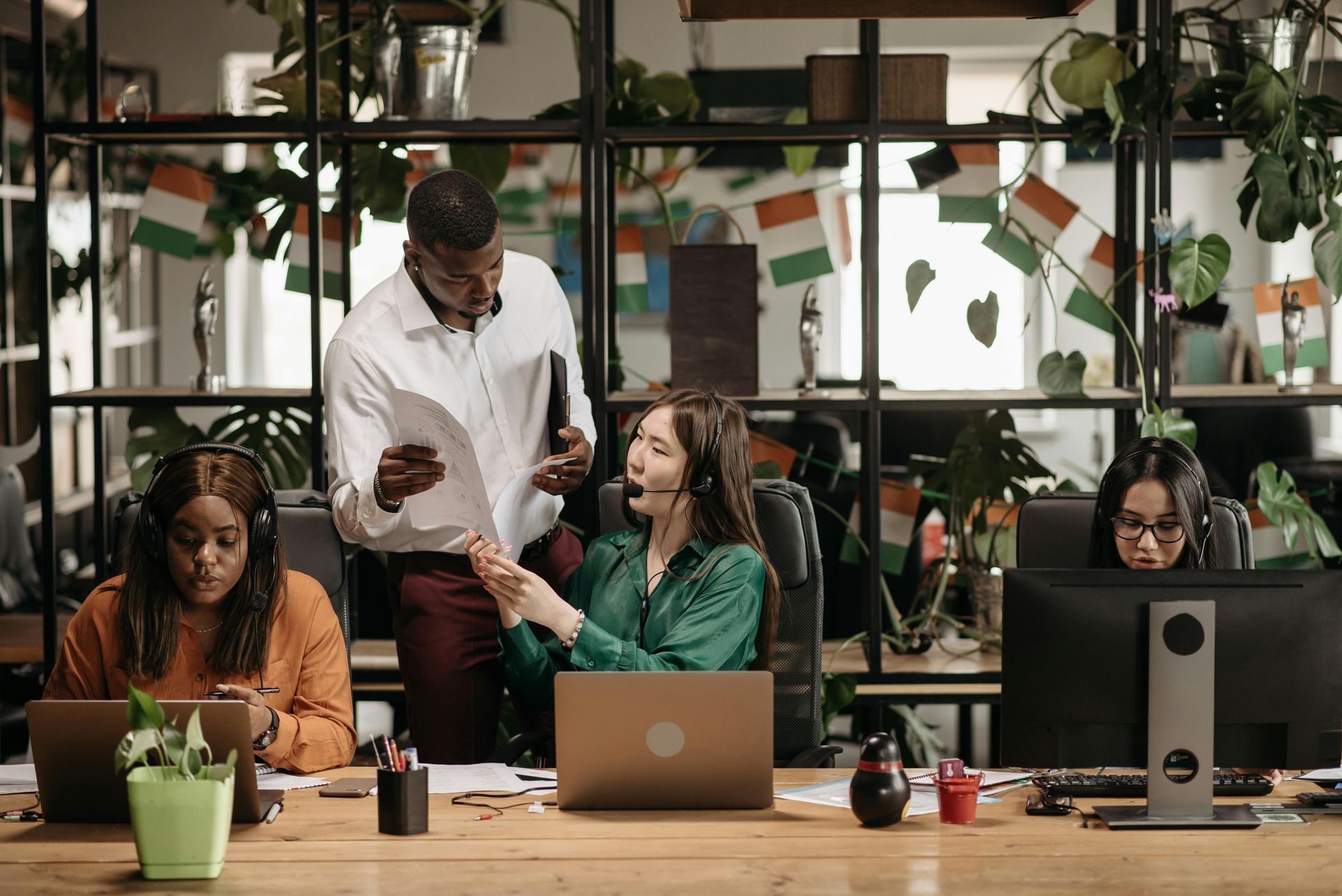 Coworkers collaborating at desks in a modern office, with laptops and a standing discussion.