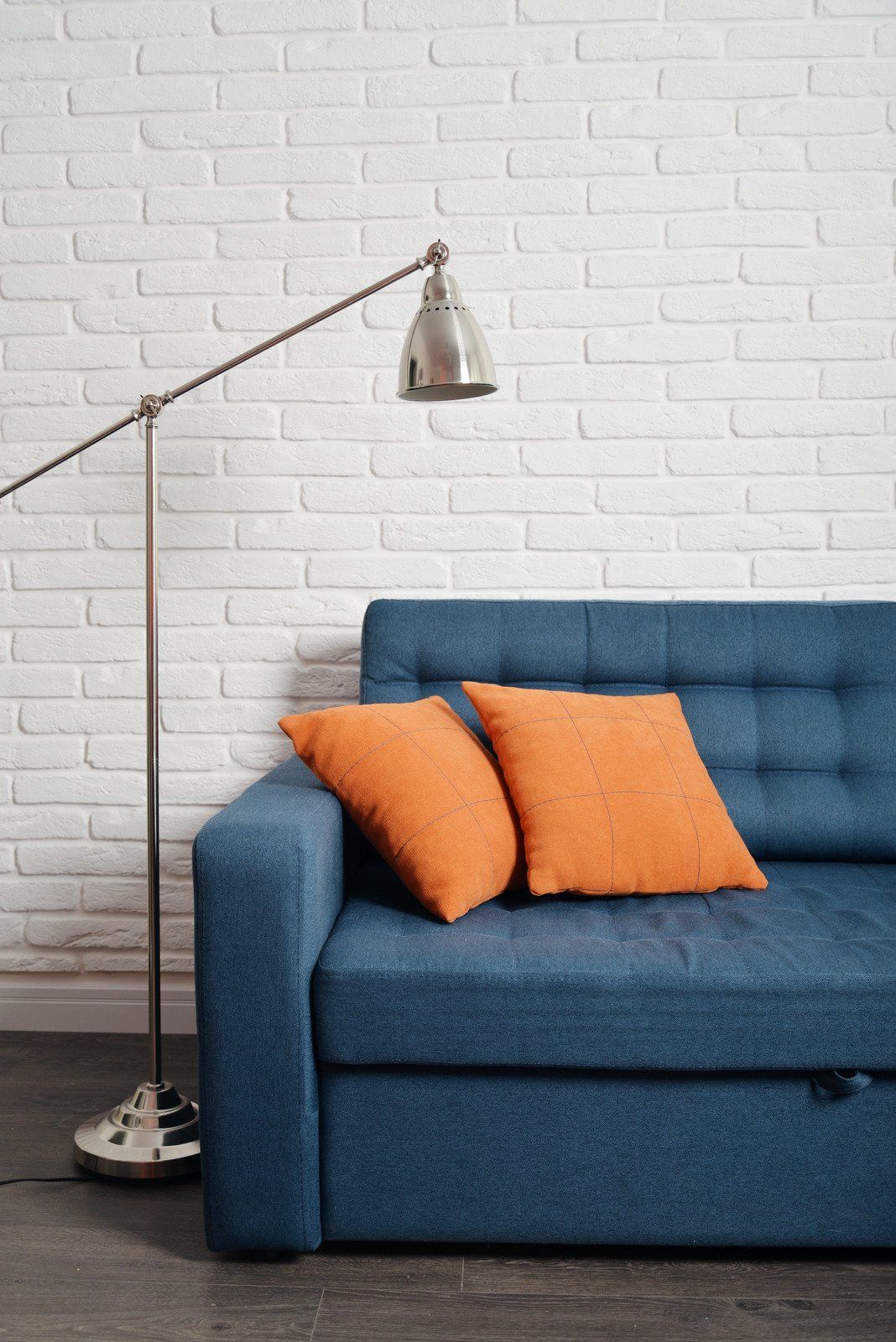 A blue couch with orange pillows and a lamp in front of a white brick wall.