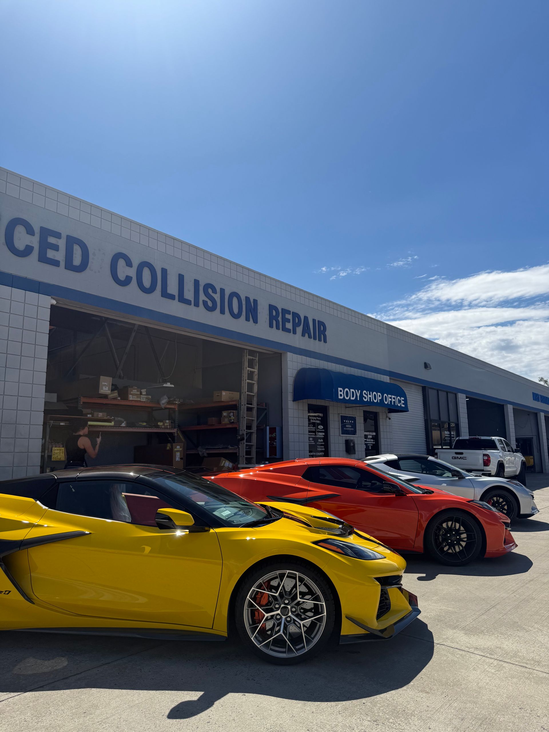 Two sports cars, yellow and red, parked outside a collision repair shop under a blue sky