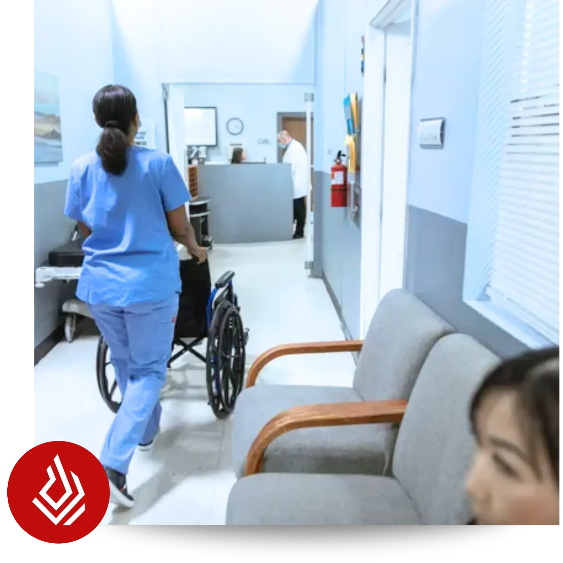 A healthcare professional in blue scrubs pushing an empty wheelchair down a bright clinical corridor.