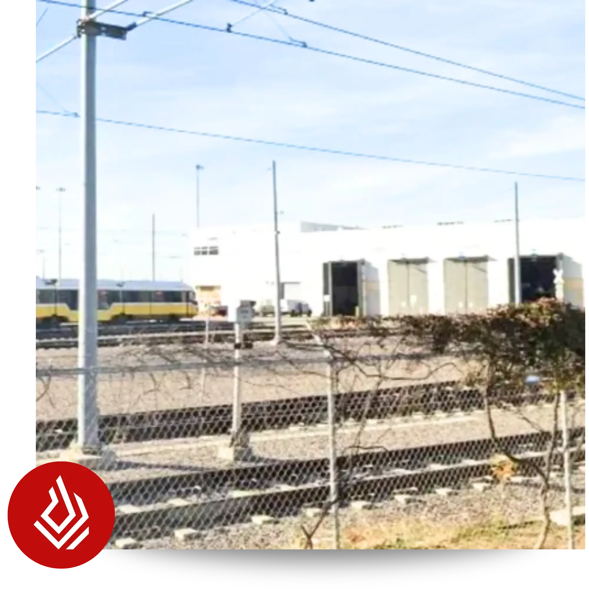 View of train tracks and a yellow train car in front of a white industrial warehouse behind a chain-link fence