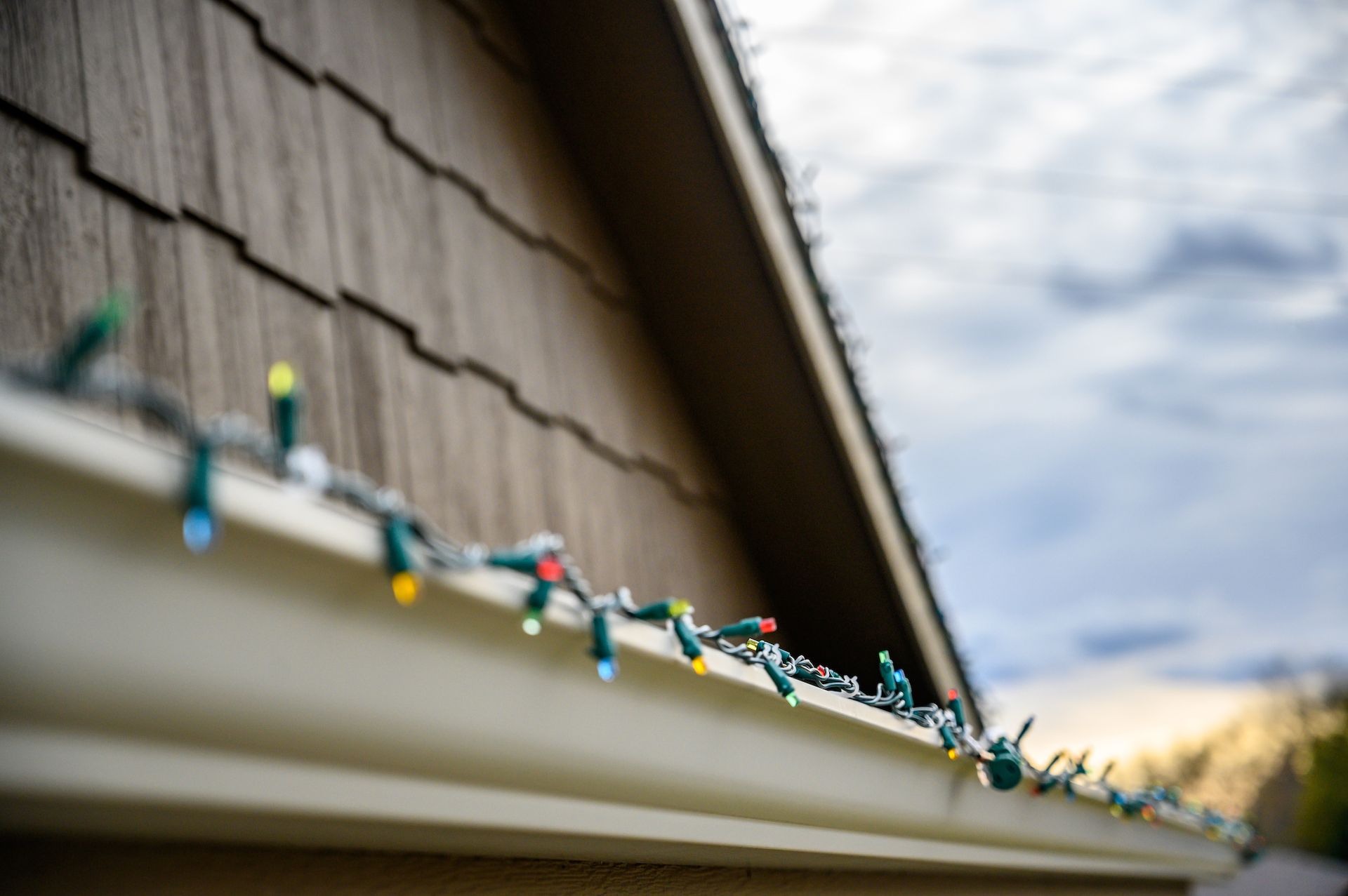 Christmas lights strung along a gutter on a brown-shingled house, with a cloudy sky in the background.