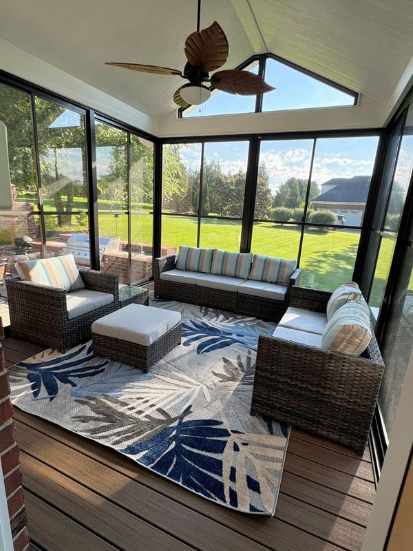 Sunroom with wicker seating, blue leaf rug, and large windows overlooking a sunny lawn and lake.
