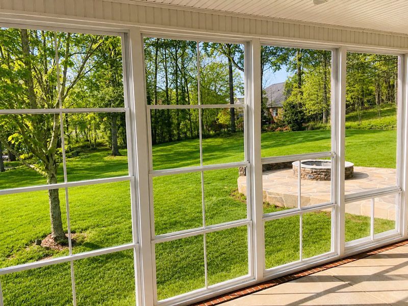 Sunny backyard seen through large white-framed windows, with green lawn, trees, and a patio fire pit.