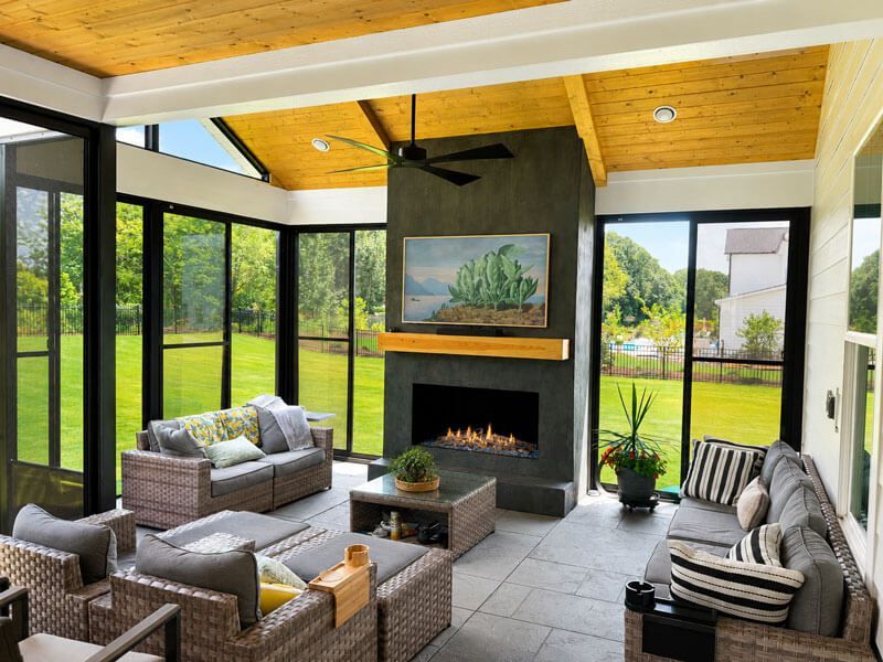 Modern screened porch with fireplace, gray wicker seating, wood ceiling, and garden view