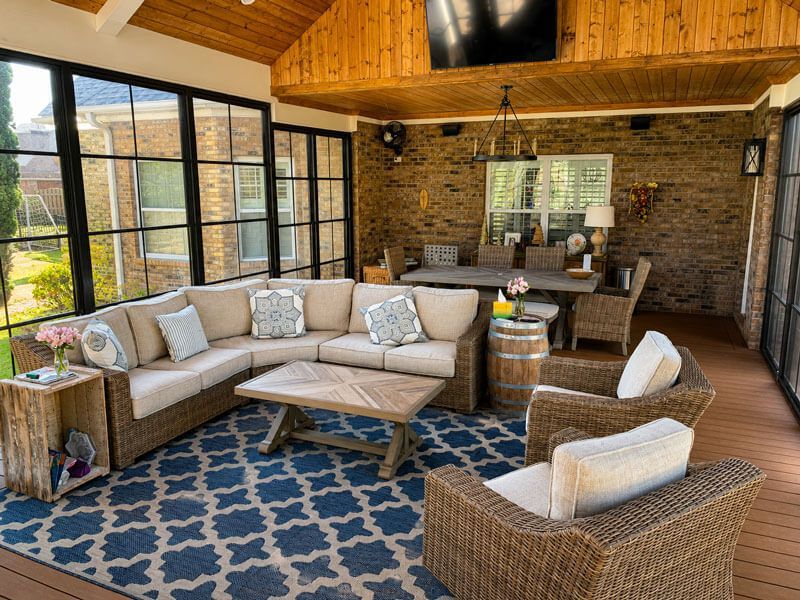 Cozy sunroom with beige sectional, woven chairs, blue rug, brick wall, and wood ceiling