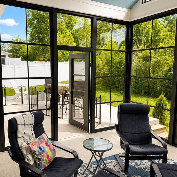 Sunlit glass-paneled porch with black chairs, glass table, and lush green yard outside.