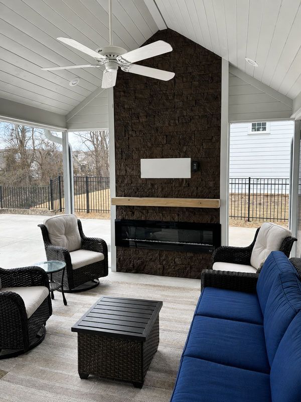 Covered patio with blue sofa, black chairs, wicker table, and stone fireplace under a vaulted ceiling.